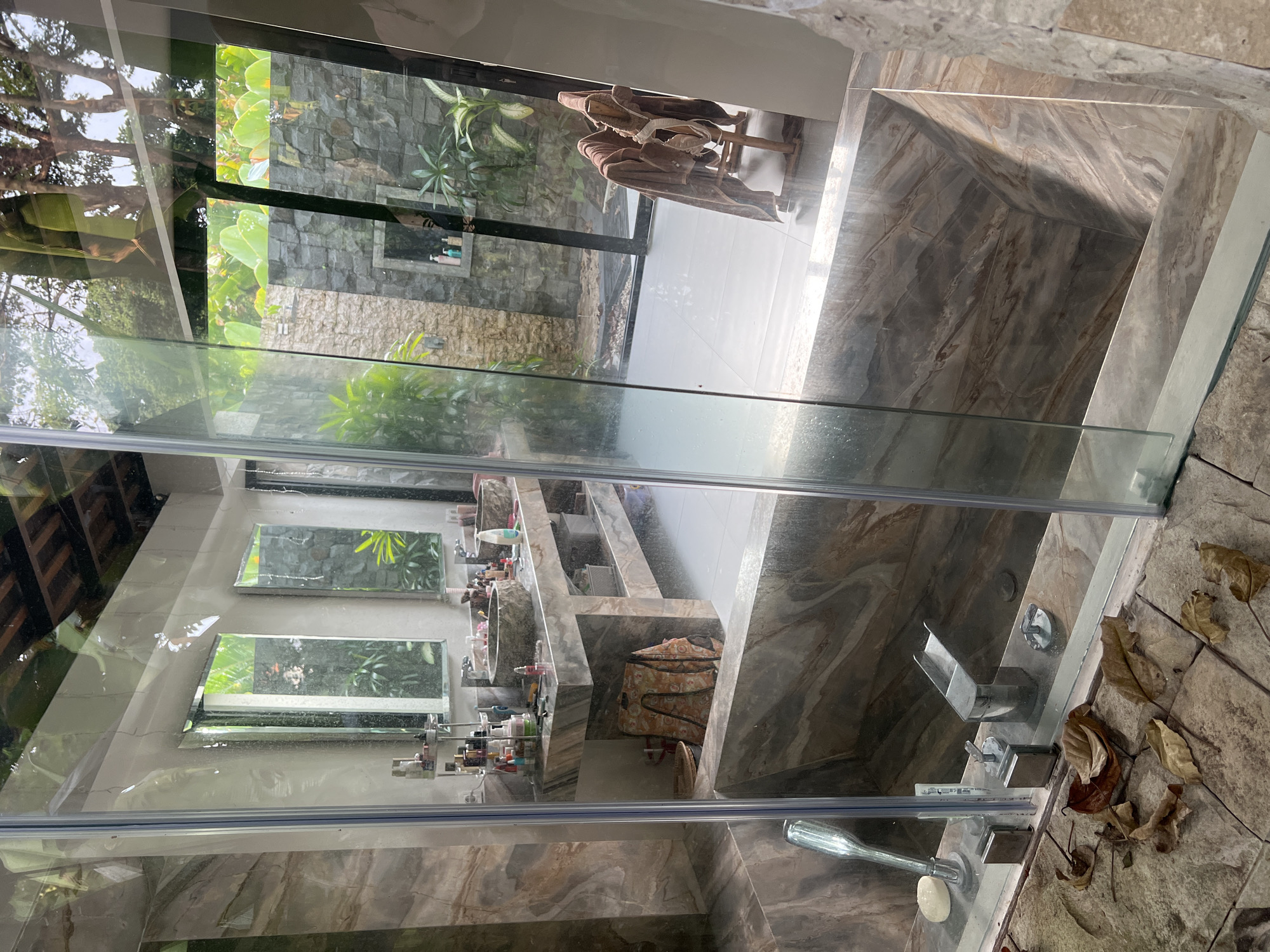 The image depicts the interior of a bathroom in Kerobokan, Indonesia, as viewed through a glass shower partition. The bathroom features marble-like tiling. Through the glass, one can see a reflection of a vanity area with multiple shelves holding various toiletries. Windows above the vanity reflect an exterior view of lush greenery. Beyond the shower, an outdoor space is visible, containing plants, a stone wall, and a clothes rack with clothing. The surrounding area appears to be a garden. The flooring outside the shower area is stone-tiled, with some fallen leaves scattered on the surface. The shower fixture, including the faucet and showerhead, are visible.