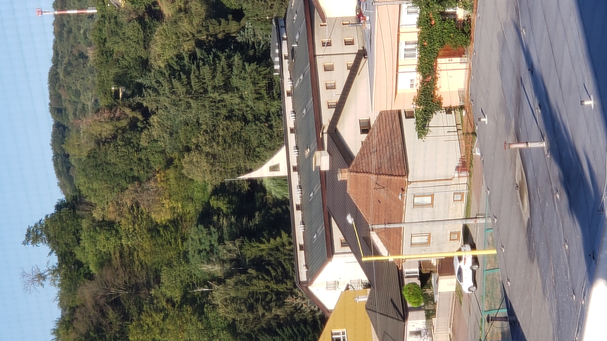 This image captures a view of a residential area nestled against a forested hillside. In the foreground, a flat, dark gray rooftop with several ventilation pipes and small protrusions is visible. Beyond this, a street level view reveals several buildings with tiled roofs. One prominent building features a reddish-brown tiled roof and light-colored walls. To its left, a yellow building stands, and further back, a larger, light-colored building with a dark gray roof and numerous windows is visible, topped by a distinctive triangular architectural element, possibly part of a church or similar structure. A white car is parked on the street in front of some of these buildings. The background is dominated by a dense green forest covering a steep hillside, with a tall, red and white striped communication tower visible on the far right of the hill. The sky is a clear, bright blue. The scene appears to be in a suburban or semi-rural part of the city of Prešov, Slovakia, given the presence of residential buildings and the natural landscape.