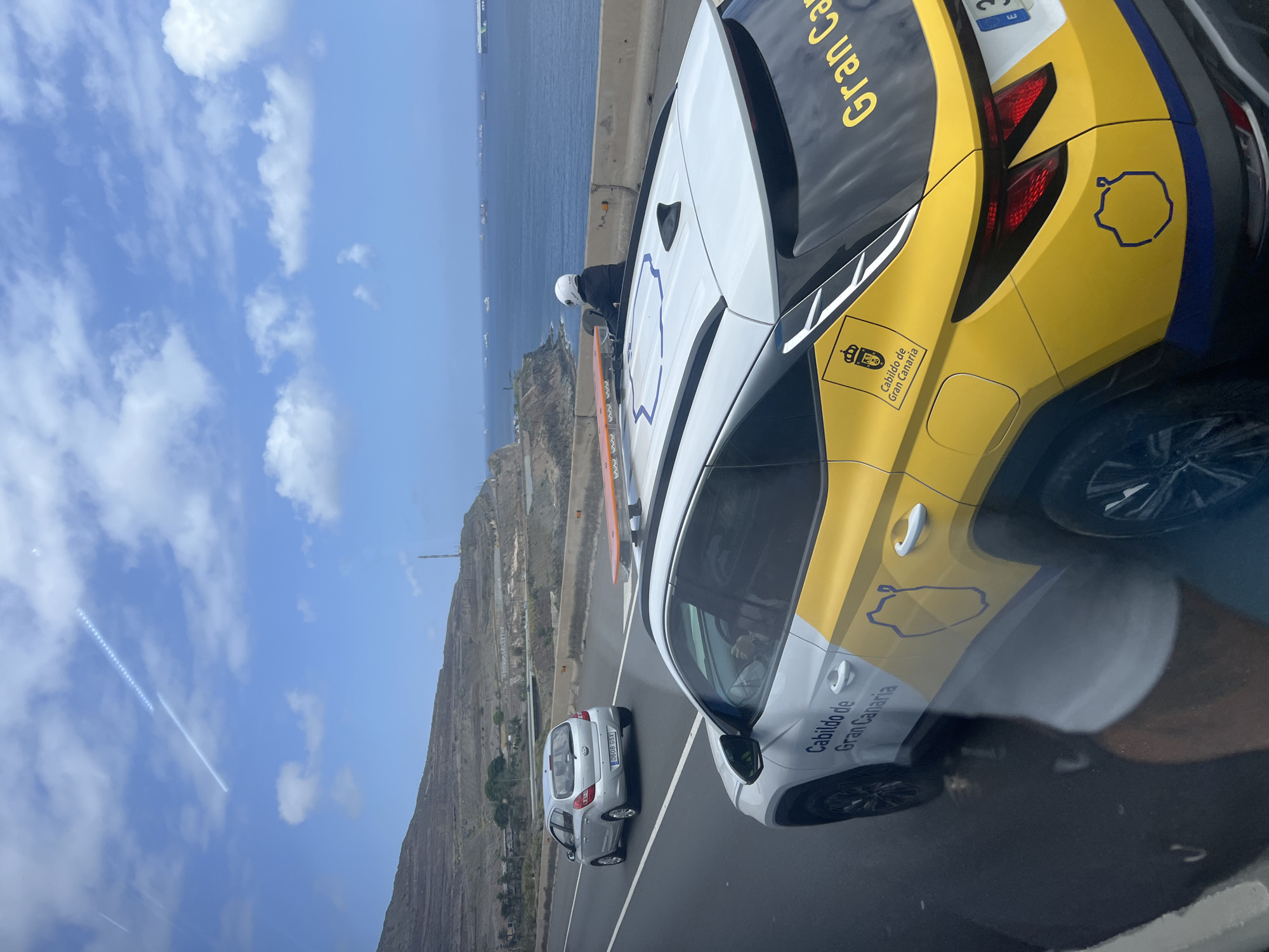 This image captures a bright daytime scene on a coastal highway, likely taken from inside a moving vehicle due to reflections in the foreground. The primary focus is a distinct white and yellow SUV-type vehicle, prominently displaying official branding. On its side and rear, the text "Cabildo de Gran Canaria" and "Gran Can" are clearly visible, along with an emblem resembling a coat of arms and an outline of an island, strongly identifying the location as Gran Canaria, Spain, which aligns with the provided context of Las Palmas, Spain.
A person wearing a white helmet and dark attire is leaning over the rear of this official vehicle, suggesting involvement in a roadside activity or assistance. An orange object, possibly a traffic cone or safety marker, is positioned on the road next to the vehicle, indicating caution. Further down the road, a silver hatchback car with the license plate "0608 GSX" is also visible.
The background features a dramatic landscape: a vast blue sea with several ships dotted on the horizon, bordered by rugged, rocky cliffs and hills. The sky is clear and blue with scattered white clouds. The overall scene suggests a public service operation or roadside event unfolding against a scenic, sunny, coastal backdrop.