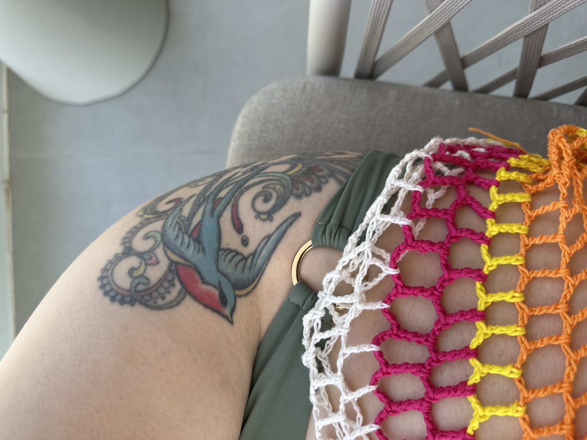 The image depicts the upper thigh and hip of a light-skinned person with a visible tattoo of a blue and red bird surrounded by swirls and floral-like patterns. The person is wearing a dark green swimsuit with a gold ring detail. A multi-colored crocheted mesh garment is draped over the thigh, featuring white, pink, yellow, and orange yarn. The background shows a light gray surface and the back of a light gray chair. The location is unspecified, the city of Choeng Thale, Thailand is only a contextual guide.