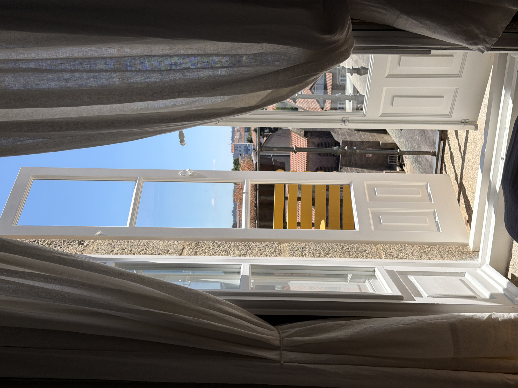 The image shows an open balcony door with sheer curtains framing the view of a cityscape under a clear blue sky. The doorway is white, with two panels on the lower half and a large glass pane above, allowing sunlight to stream in. The walls are made of rough, light-colored stone. Outside, red-tiled roofs and buildings are visible. A yellow slatted balcony railing is prominent in the middle ground. The setting is likely a room in a building in Santiago de Compostela, Spain, given the architectural style and red-tiled roofs common in the region. No people are visible in the scene.