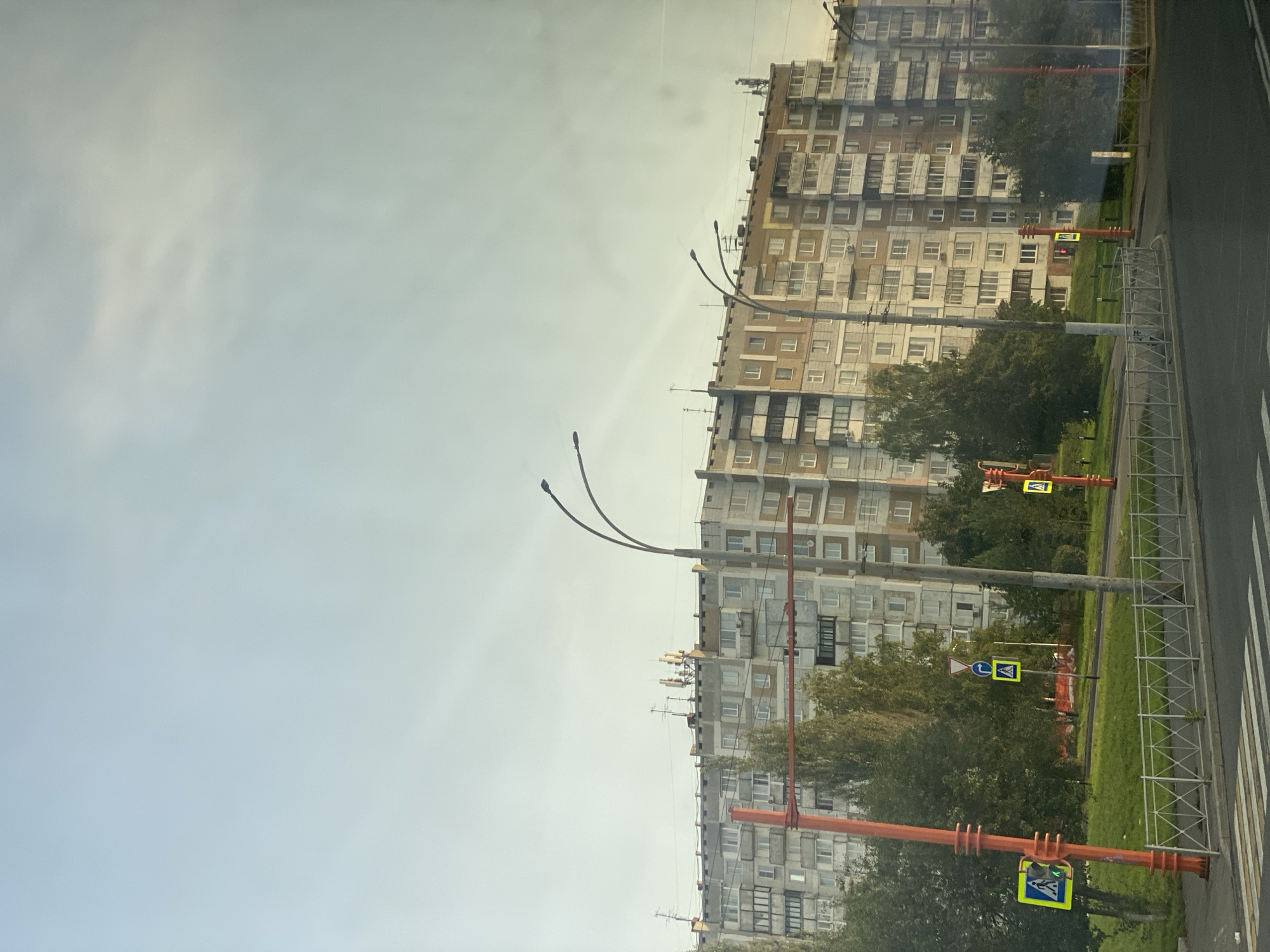 The image captures a view of a multi-story residential building in Kemerovo, Russia, under a pale, overcast sky. The building, with its numerous balconies and windows, appears to be constructed in a Soviet-era style. In the foreground, a street with a pedestrian crossing is visible, marked by white stripes. A red traffic signal pole with a pedestrian crossing sign is positioned to the left. Various traffic signs, including directional and pedestrian crossing signs, are present along the roadside. Trees and a grassy area separate the street from the building. Streetlights with curved arms are visible over the scene. The overall impression is of a typical urban landscape.