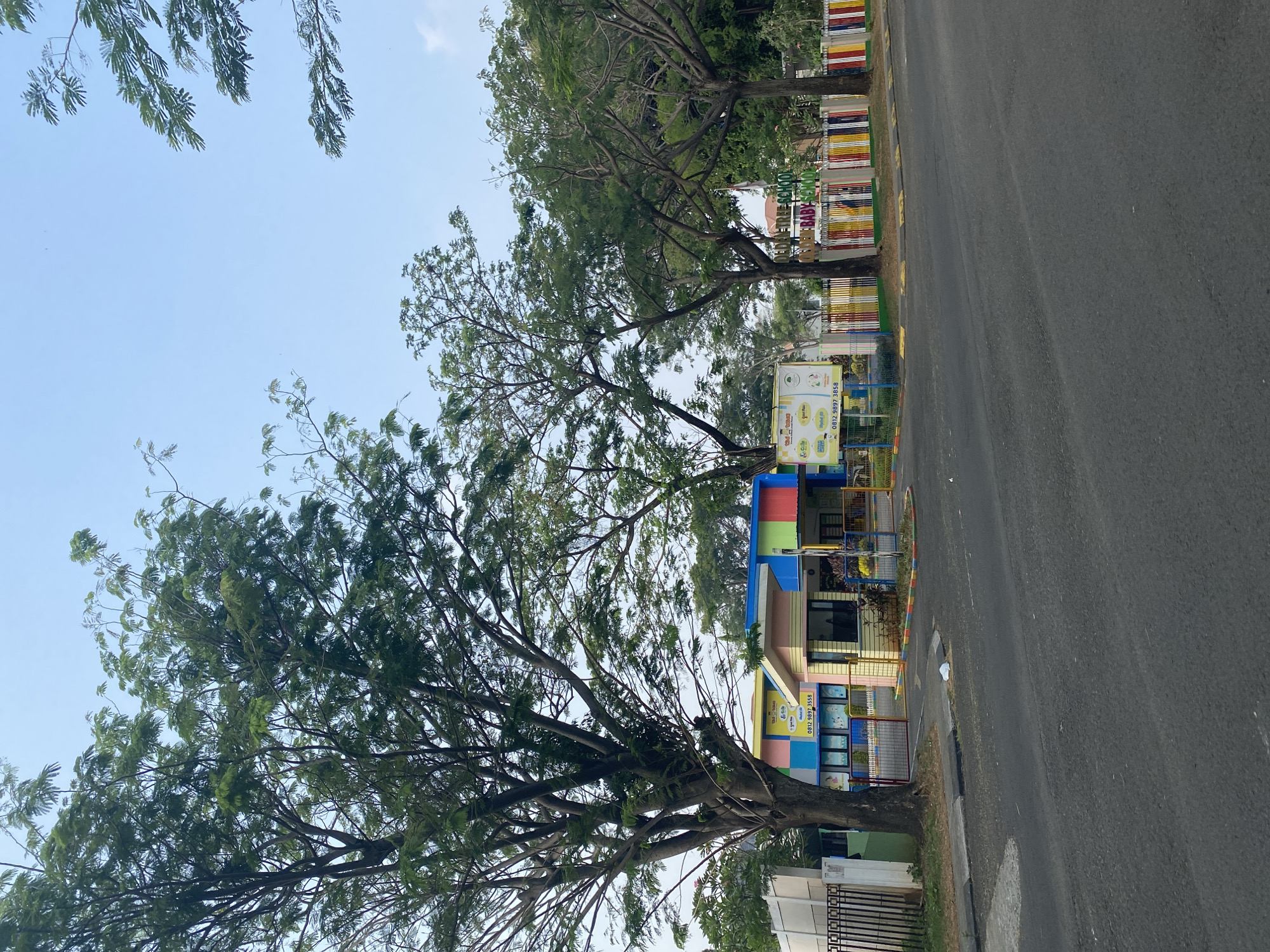 The image depicts the exterior of a brightly colored school building, likely a kindergarten or preschool, situated in East Jakarta, Indonesia. The building features a vibrant facade with sections painted in yellow, pink, blue, and green. A security booth is positioned at the entrance, which is flanked by a colorful fence adorned with red, yellow, blue, and white vertical bars. Large trees with lush green foliage are scattered throughout the scene, providing shade and framing the school. A paved road runs in front of the school, with a sidewalk separating it from the property. The sky above is a clear, pale blue. There are no people visible in the image. The overall impression is that of a well-maintained educational facility set against a backdrop of natural greenery.