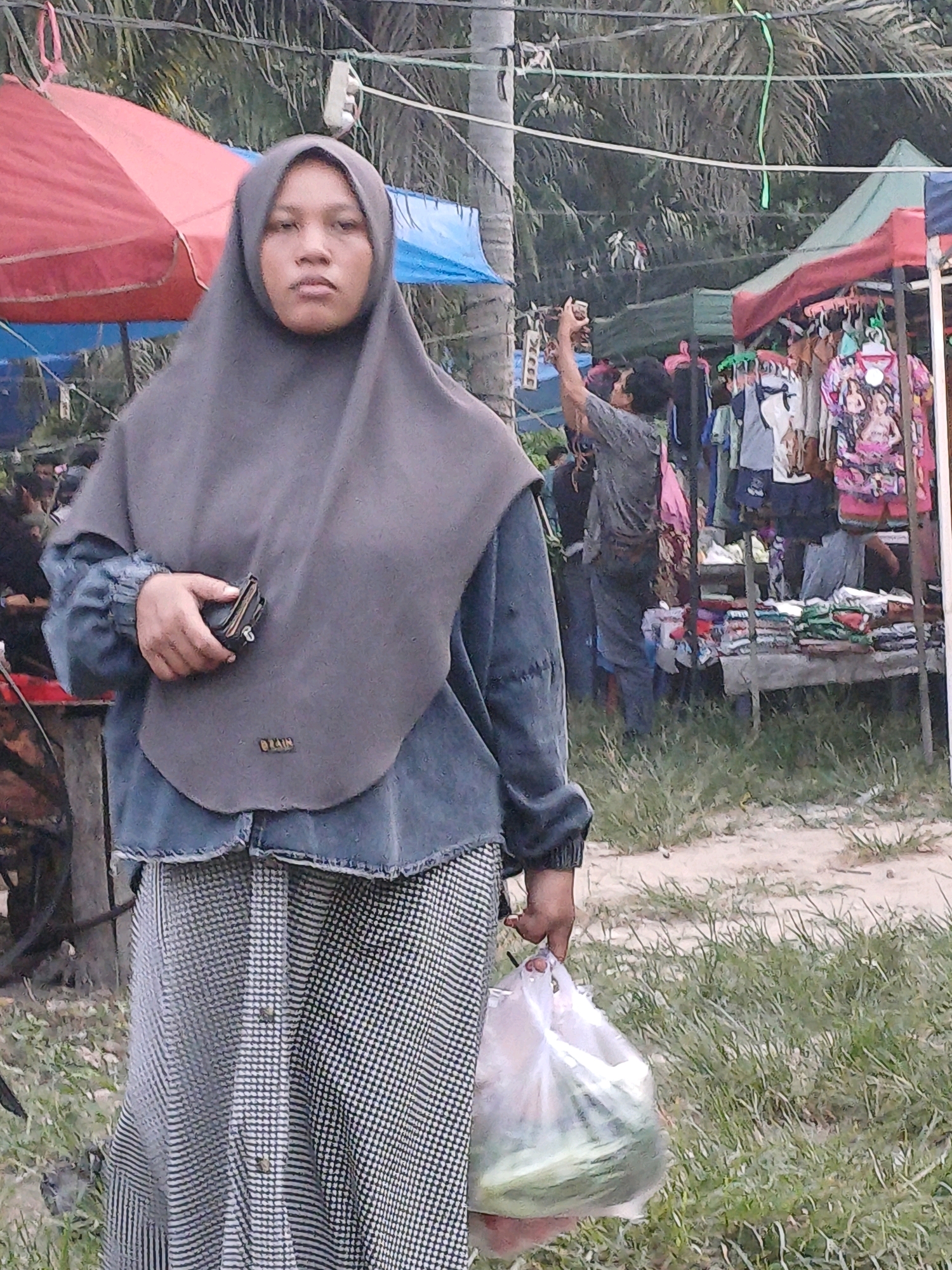 An overhead shot captures a young woman, likely in her late teens or early twenties, wearing a gray hijab and a blue denim-like jacket over a black and white checkered top. Her left hand clutches a black wallet. She is holding a clear plastic bag filled with green vegetables in her right hand. The background reveals a bustling outdoor market scene. A red and blue canopy stretches across the upper left, possibly indicating a stall or shelter. In the middle ground, a man in a gray t-shirt and dark pants stands, holding a phone up to take a picture, with other market stalls and merchandise visible behind him. The ground is covered with sparse vegetation and sand. The image appears to be taken in the city of Dumai, Indonesia.