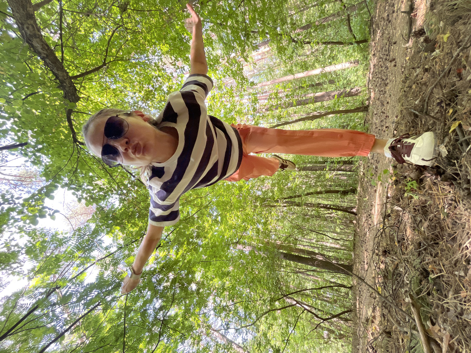 A woman stands in a forest in the city of Chaikovsky, Russia. She wears sunglasses, a black and white striped t-shirt, and orange pants. Her arms are outstretched. One of her legs is bent, revealing her brown and white sneaker. The background includes dense trees and foliage.