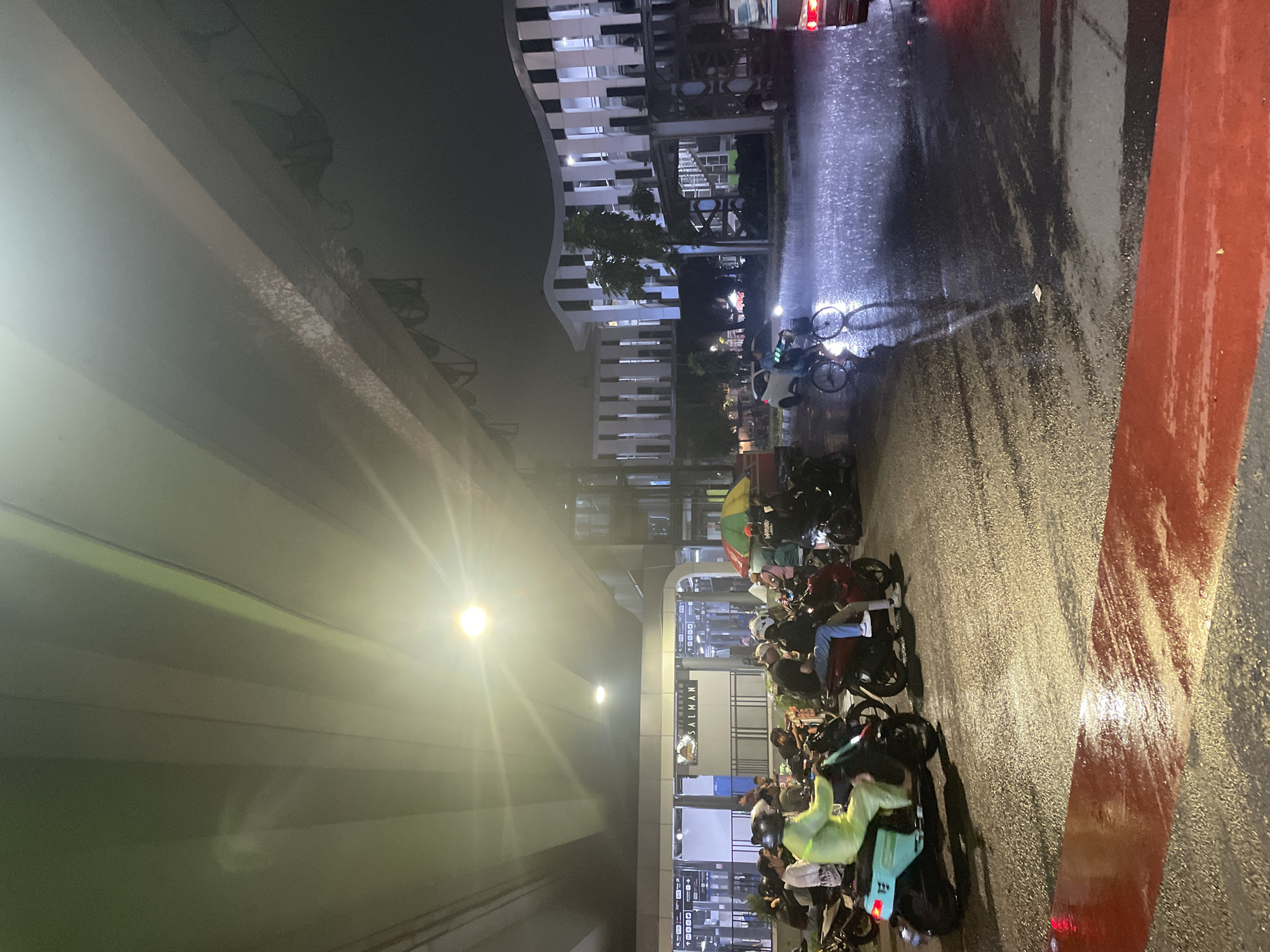 The image depicts a wet street scene at night in Central Jakarta, Indonesia. The perspective is angled, showing an elevated concrete structure above, illuminated by lights. A row of parked motorcycles lines the street, many occupied by people, likely waiting. Some individuals are wearing helmets and rain gear. In the background, there is a modern building with illuminated windows. The road surface is wet and reflective, suggesting recent rainfall. A broad red stripe is visible on the right side of the road. The signage "Stasiun Sudirman" (Sudirman Station) is visible.