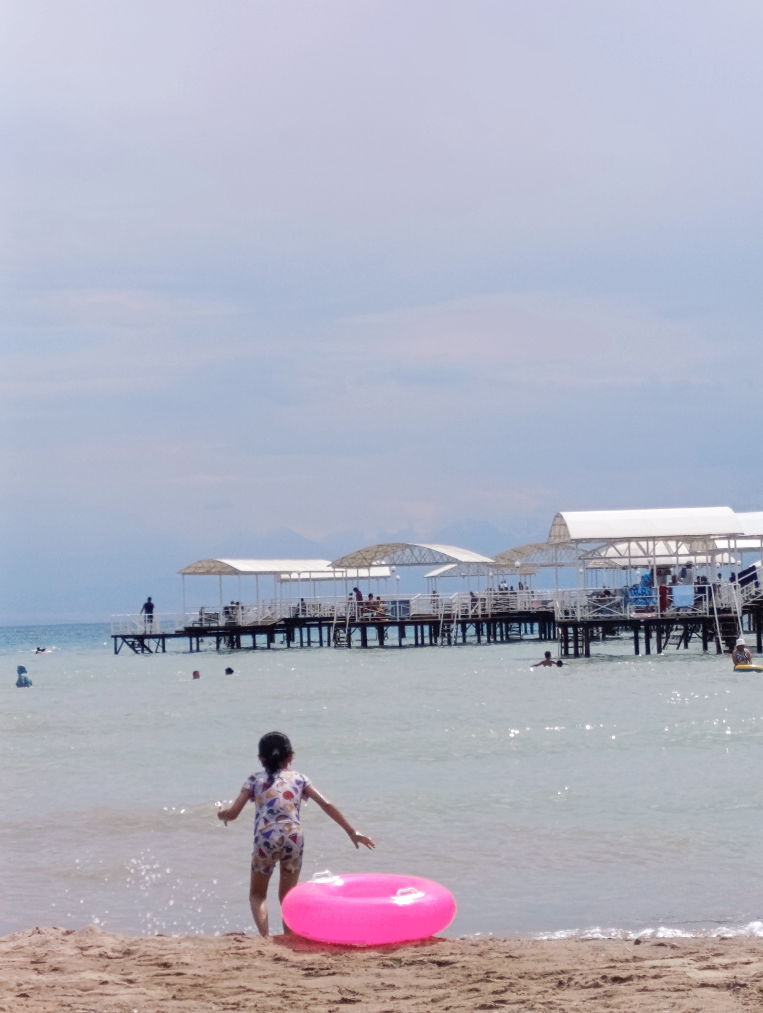The image depicts a beach scene at Sary-Oy, Kyrgyzstan, under a bright, overcast sky, suggesting it's daytime. The foreground features a sandy beach with a young girl in a colorful swimsuit standing near the water's edge. She has a bright pink inflatable ring placed beside her on the sand.
In the background, a wooden pier extends into the calm, light blue water. The pier is lined with white canopies, providing shade, and there are people visible on and around it, some swimming in the water and others relaxing on the pier. The overall atmosphere appears relaxed and summery, typical of a beach resort. The distant horizon shows faint outlines of mountains, hinting at the geographical setting. The water is relatively clear, with some ripples and a few people swimming further out. The overall impression is one of a peaceful day at a popular tourist destination. No text is visible in the image.