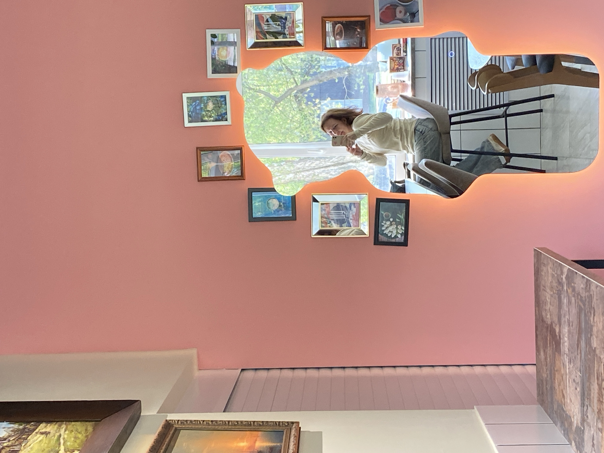 A woman is taking a selfie in a mirror in Chaikovsky, Russia. The mirror is bordered with small framed images and illuminated with an orange light. The room has pink walls, and there are other framed pictures on the wall below. The reflection in the mirror shows a window with a view of trees, a bar with chairs, and a wall with vertical stripes.