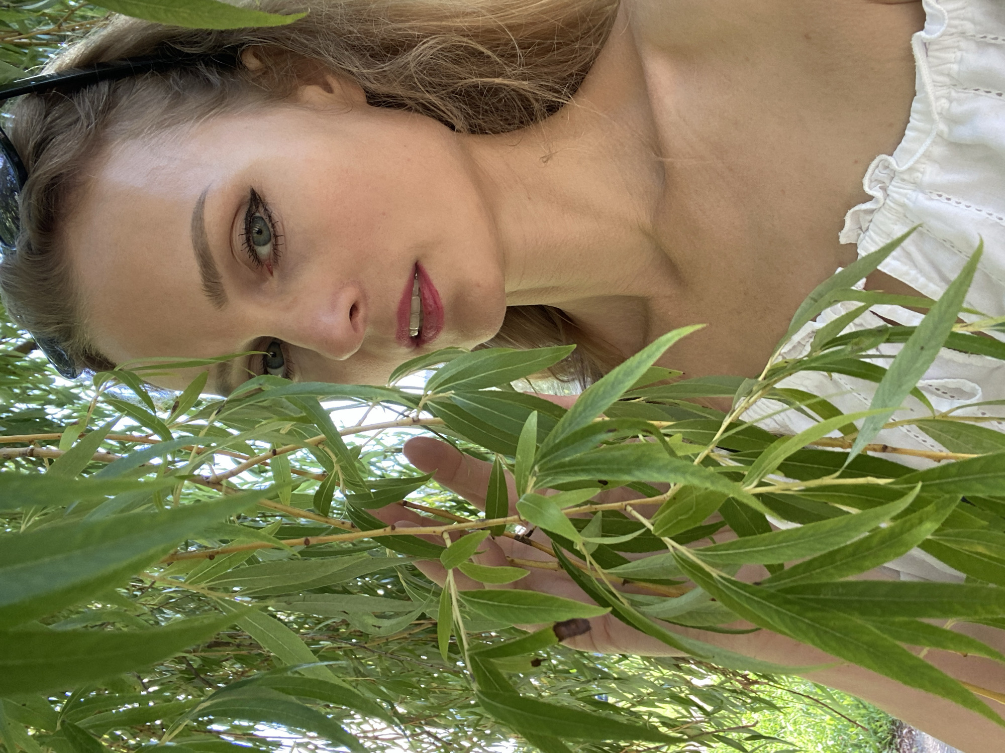 A young blonde woman is positioned in the foreground, partially obscured by the drooping branches and leaves of a willow tree. She is looking towards the camera with a slight smile. She is wearing a white, off-the-shoulder top with delicate ruffles and eyelet detailing. Her makeup includes winged eyeliner and pink lipstick. Sunglasses are pushed up onto the back of her head. Her right hand is visible, gently touching a branch. The background shows glimpses of greenery and sunlight, suggesting an outdoor setting. The image does not contain any specific visual cues that would allow for identification of the location within the city of Lida, Belarus.