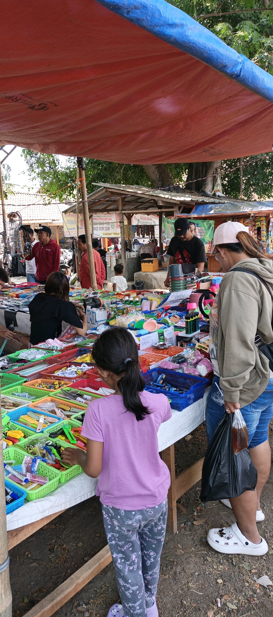 In Jatinom, Indonesia, a bustling outdoor market scene unfolds under a large, red and blue tarp. The market is filled with various goods displayed on tables and stalls. In the foreground, a young girl with dark hair pulled back in a ponytail, wearing a pink t-shirt and patterned leggings, examines items in a green plastic basket. She is standing on the ground, looking down at a table overflowing with colorful stationery and small tools. To her right, a woman with dark hair, wearing a light brown hoodie and blue shorts, stands with her back to the camera, holding a black plastic bag containing a dark liquid. She is wearing white clogs. Behind them, other market-goers are visible, including a man in a black t-shirt and baseball cap, and a woman with dark hair wearing a black shirt. Further back, a child in a white shirt is visible near a wooden stall. The market appears to be well-stocked, with various plastic baskets filled with an assortment of products, including what appear to be pens, knives, and other small accessories. The ground beneath the stalls is dirt and leaves, indicating an open-air setting. The overall atmosphere suggests a lively marketplace with people browsing and shopping.