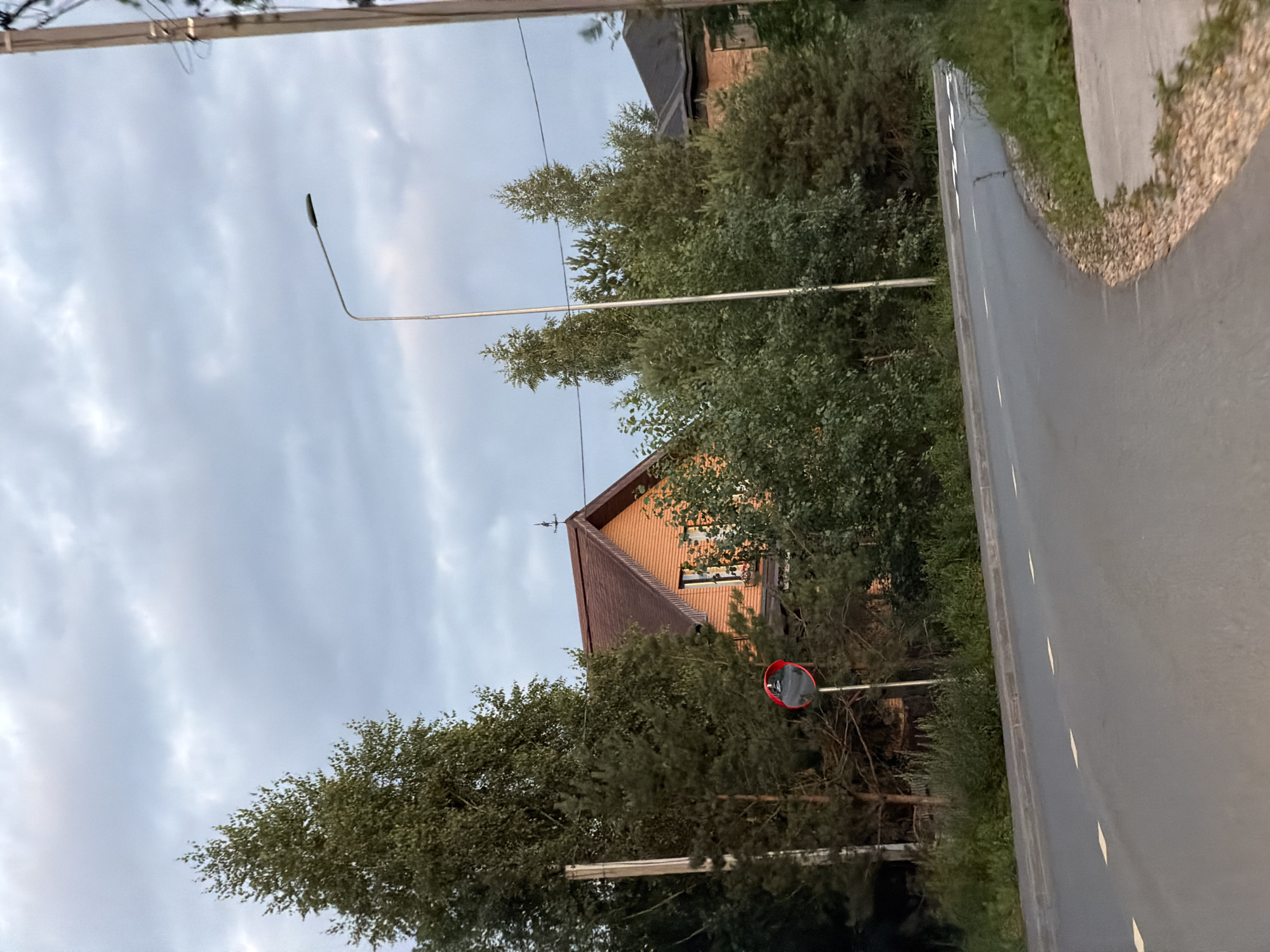 The image captures a rural road in Kazan, Russia, under a cloudy sky, suggesting it is either early morning or late afternoon. The road, paved and marked with white dashed lines, curves to the right, bordered by gravel and lush green vegetation. On the left side of the road, a tall street lamp stands, its light fixture angled downwards. Further back, a house with a brown tiled roof is partially visible, nestled among dense trees. A red and black convex mirror, commonly used to improve visibility on blind corners, is mounted on a pole near the road. The overall scene conveys a quiet, serene atmosphere typical of a rural setting in Russia. There is no visible text in the image.