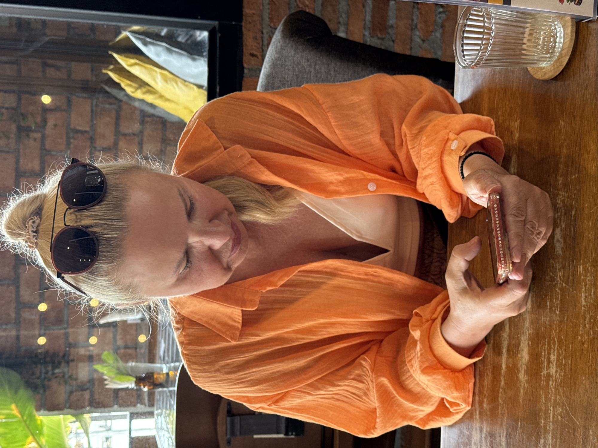 A blonde woman wearing an orange shirt and sunglasses on her head sits at a wooden table, looking at her phone. She has her hair in a messy bun. In the background, there is a brick wall, a window with plants and lights, and a dark chair. A glass with vertical lines is on the table next to her. The scene appears to be in a restaurant or cafe in Ko Samui District, Thailand.