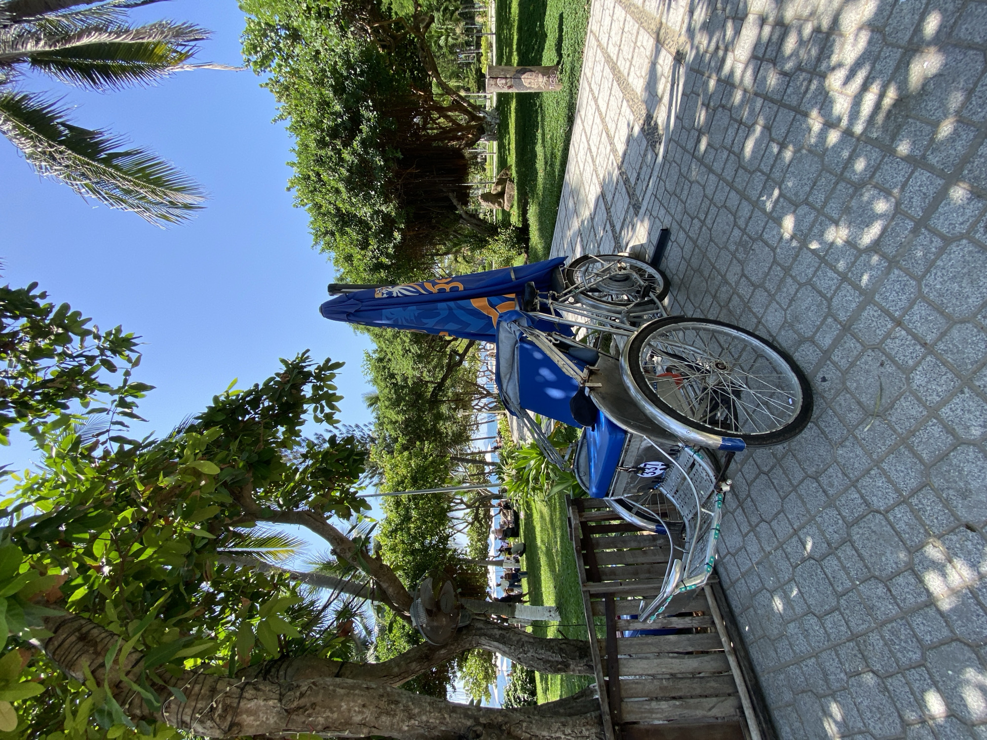 A pedicab with a blue seat and a folded blue umbrella stands on a cobblestone path in Nha Trang, Vietnam. The pedicab is parked near a wooden fence and under the shade of a large tree. In the background, lush green trees line a park area with people relaxing on the grass and near the beach. The sky is a clear blue, indicating a sunny day. The scene depicts a leisurely outdoor setting, likely a tourist area in Nha Trang.