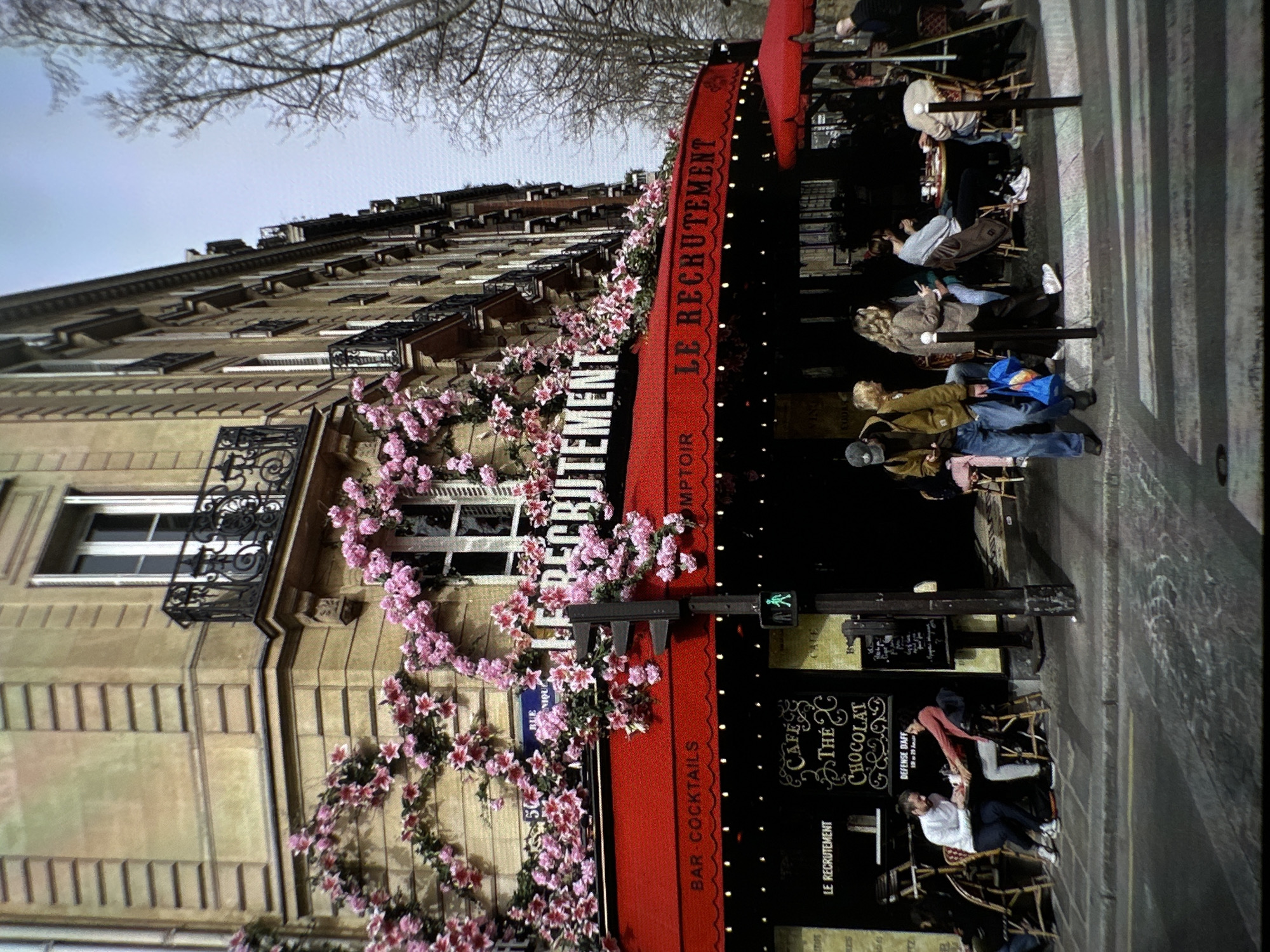 Here's a detailed description of the image:
**Scene:** A street scene in Paris, France, featuring a building with a café called "Le Recrutement". The building has a classic Parisian architectural style with multiple floors, windows with ornate iron balconies, and a beige facade.
**Objects:**
* **Flowers:** Pink flowers are draped across the building's facade.
* **Awning:** The café has a red awning with decorative lights.
* **Signage:** The awning has the name "Le Recrutement" printed on it. There is a chalkboard menu outside the café advertising "Café, Thé, Chocolat".
* **Tables and chairs:** Tables and chairs are set up outside the café for customers.
* **Street:** There is a pedestrian crossing in the foreground and a traffic light.
**People:**
* People are sitting at the café tables, some drinking and relaxing. Their actions are casual and suggestive of a relaxed atmosphere.
**Environment:**
* The scene takes place during the daytime.
* The street appears to be a typical Parisian street with buildings on either side.
* Bare tree branches are visible in the background, suggesting either late autumn or early spring.
**Location:** The image clearly shows a scene in Paris, France. There is no evidence suggesting a location in Chișinău, Moldova.