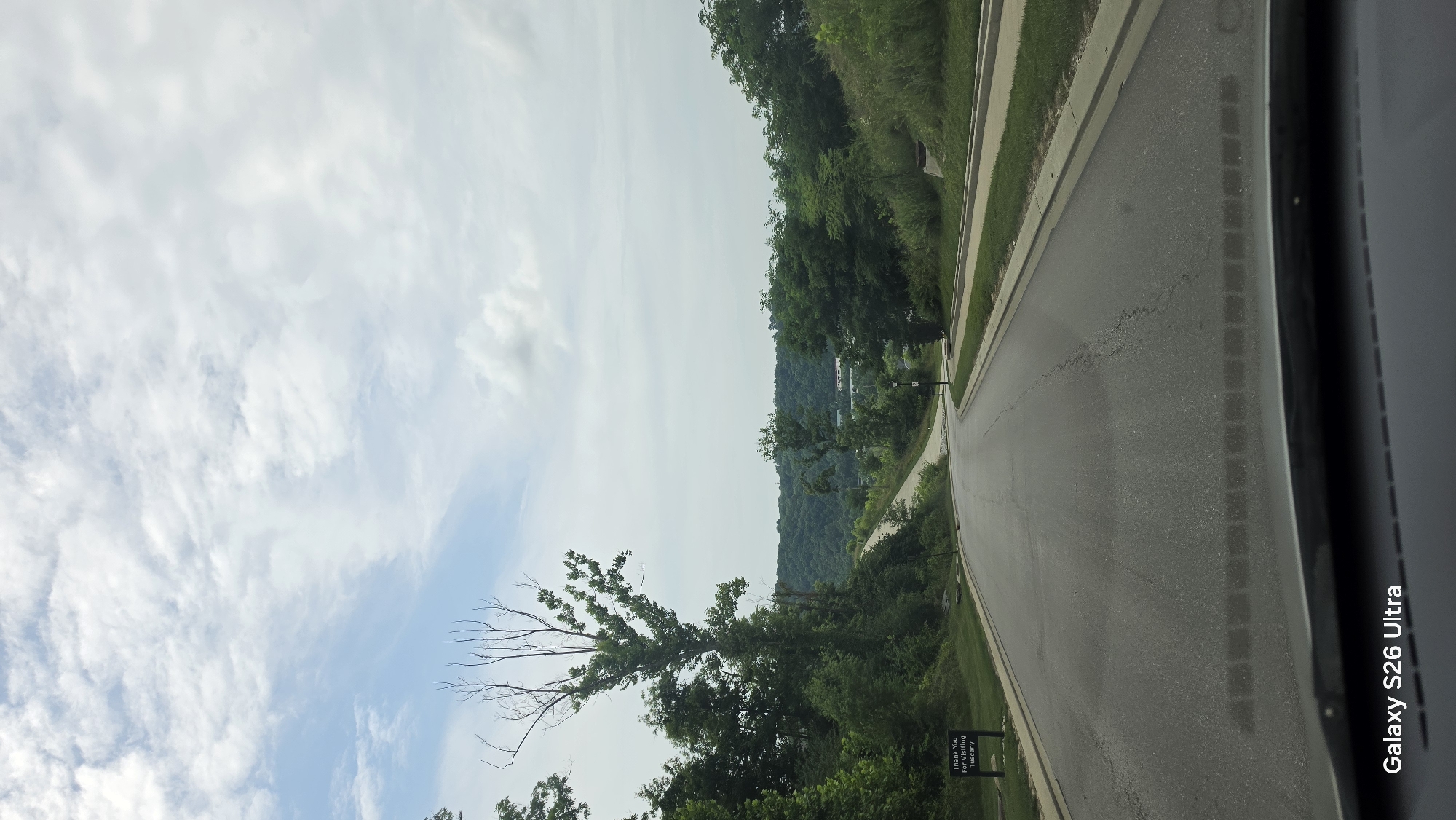 This media file shows a paved road curving through a landscape of rolling green hills and trees under a partly cloudy sky. The perspective is from inside a vehicle, looking out the side window or windshield. A concrete curb and a strip of grass run alongside the road. The trees are lush and green, indicating it is likely spring or summer. The sky is a mix of blue and white clouds, suggesting a bright but not entirely sunny day.
In the distance, more densely wooded hills are visible. On the right side of the road, a dark-colored object, possibly a signpost or a light fixture, is present. Further down the road, a dark silhouette of a sign with white text is visible. The text on this sign reads: "Thank You For Visiting Tuscany". The overall scene conveys a sense of travel or a scenic drive through a natural environment. The time of day appears to be daytime, given the lighting conditions. The image is oriented vertically, with the road and landscape running from the bottom to the top of the frame. The top of the frame is dominated by the sky with wispy white clouds. The lower portion of the frame includes the foreground of the road and the beginning of the tree line.