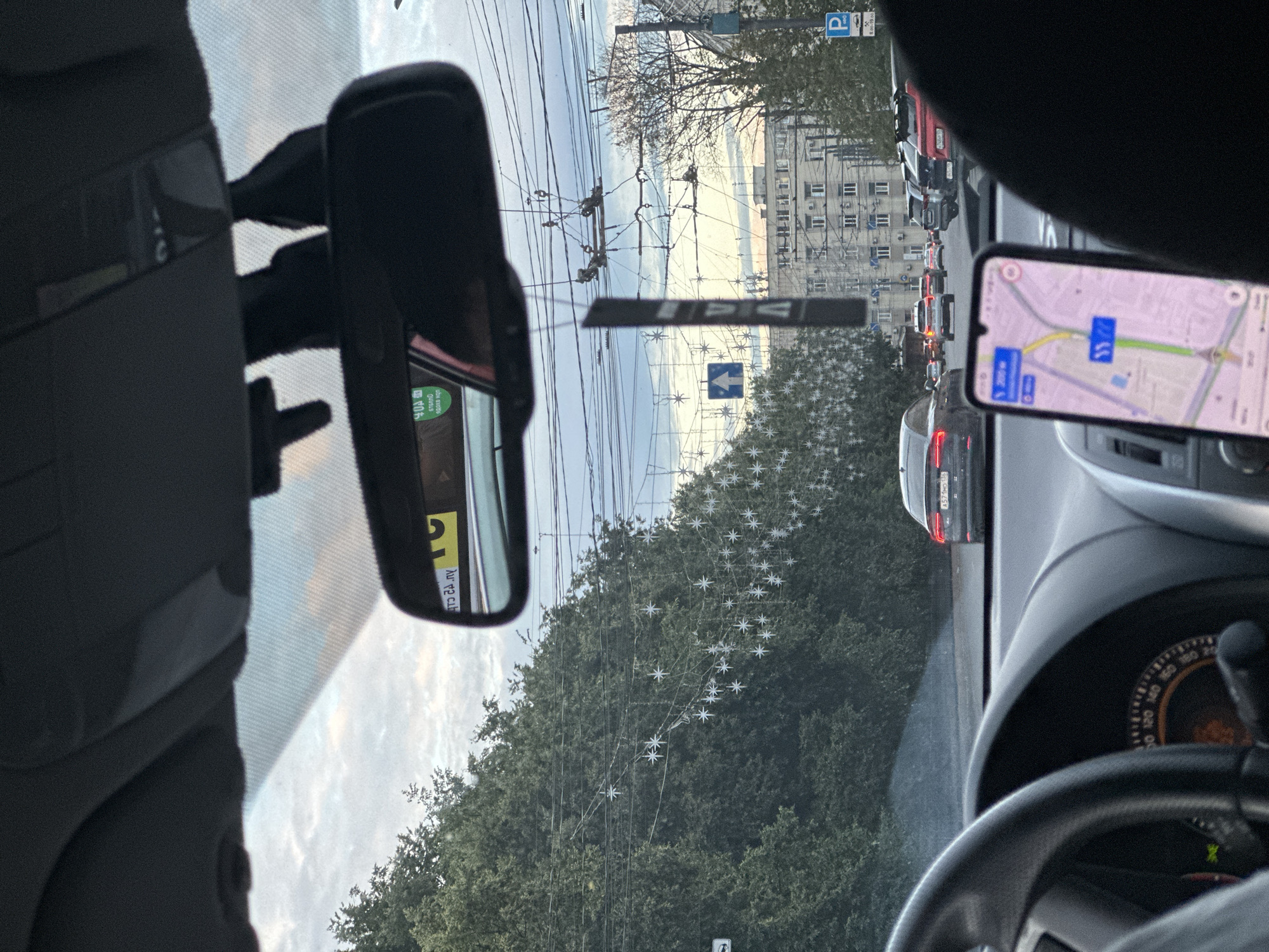 This image captures a view from inside a car, looking out through the windshield. The perspective is from the driver's seat, evidenced by the steering wheel visible in the foreground. The scene depicts a traffic jam on a road lined with trees and buildings. A smartphone mounted on the dashboard displays a navigation app, indicating the car is in motion or about to move.
The time of day appears to be late afternoon or early evening, with soft, diffused light and a pale sky. The weather seems overcast, contributing to the muted colors of the scene. The trees lining the road are dense and green, and a string of star-shaped decorations is visible, possibly indicating a festive season or holiday. In the background, several apartment buildings stand, typical of urban environments.
The traffic jam suggests a common urban commuting scenario. Various cars are visible, including a dark-colored car in the foreground and a line of vehicles stretching down the road. Overhead, power lines and trolley wires crisscross the sky, hinting at an urban setting with public transportation infrastructure.
A review mirror is also visible, reflecting a small portion of the car's interior and the sky. The overall impression is one of a typical day in a Russian city, with the driver navigating through traffic. There are no people clearly visible, but the presence of the car and the navigation app points to human activity. Text is visible on the navigation app screen, which is too blurry to read clearly, and on a sticker on the rear-view mirror, also too blurry to read.