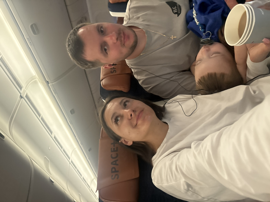 A family of three is on an airplane. A man in a gray shirt and a silver cross necklace looks at the camera. Beside him, a sleeping child with a blue hoodie lies on a woman's shoulder. The woman wears a white long-sleeved shirt and gazes upward. Airplane windows are visible on the left. Seat headrests labeled "SPACE+" are visible behind the couple. The man holds disposable cups.