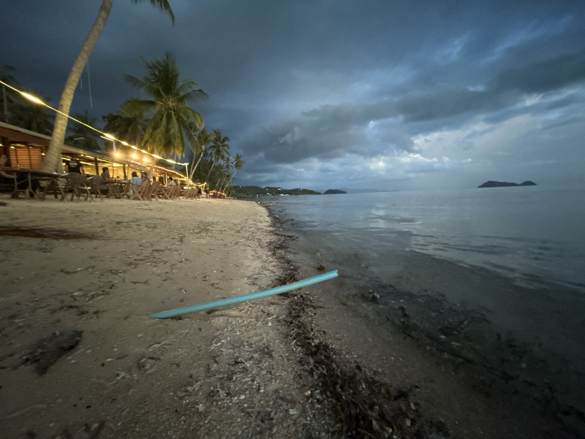 A wide shot of a beach in Koh Phangan, Thailand, at dusk. The sky is filled with dark, dramatic storm clouds. The sea is calm, with a faint silhouette of a small island in the distance. On the left side of the frame, a line of palm trees borders the beach, with string lights illuminating a row of tables and chairs where several people are seated, appearing to be at an outdoor restaurant. The sand is wet near the waterline, strewn with small pieces of debris. A long, light blue pool noodle lies on the sand in the foreground, angled towards the water.
