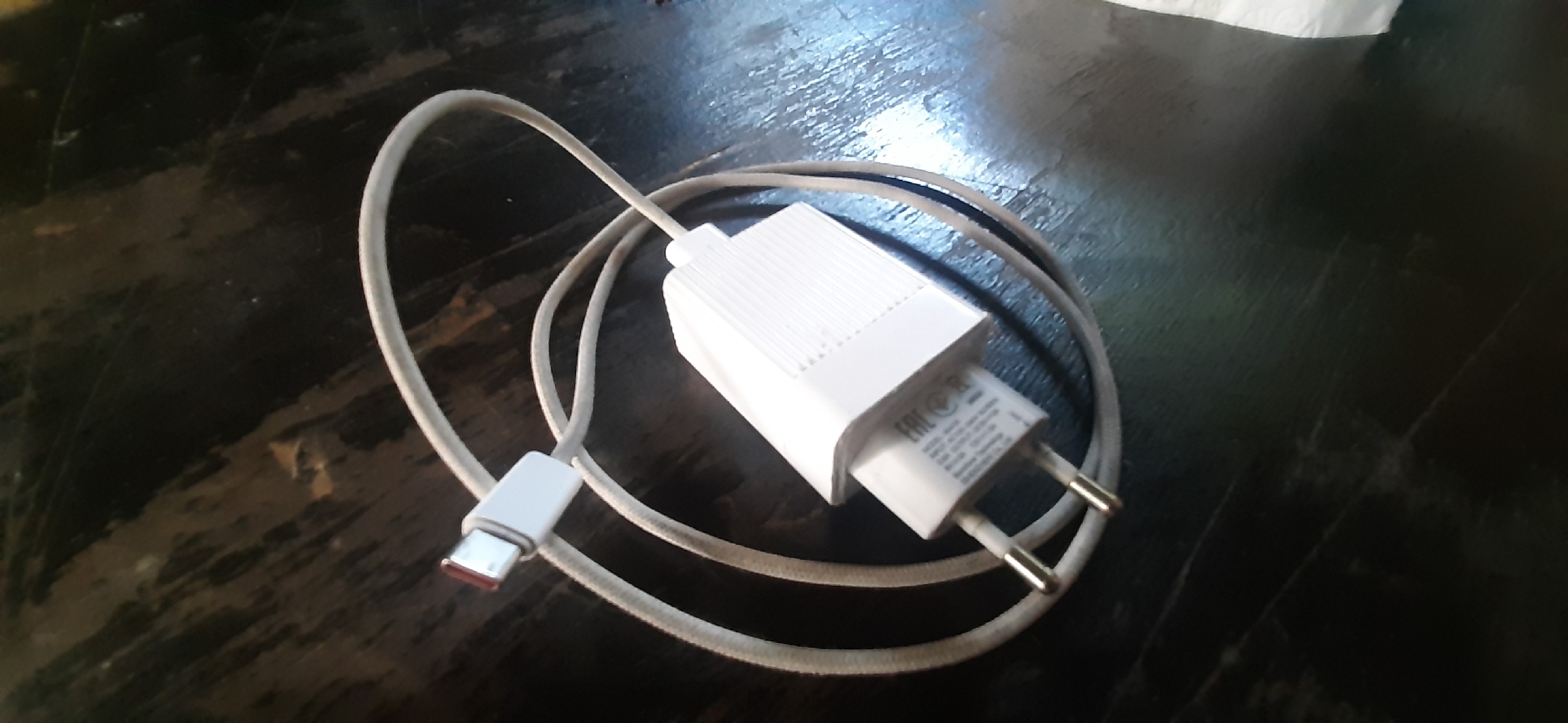 The image shows a white USB-C charger and cable resting on a dark, textured surface. The charger has two prongs for plugging into an electrical outlet and features some embossed text. The cable is coiled and has a USB-C connector on one end. The lighting suggests it is daytime. The scene appears to be indoors, possibly in a home or office, given the object and surface. There are no people visible. The surface has a wood-grain pattern with some lighter streaks and darker patches, possibly indicating wear or a deliberate finish. The overall impression is of a functional electronic accessory placed on a table or desk. Visible text on the charger includes "EAC" within a circle and other smaller print that is difficult to decipher.