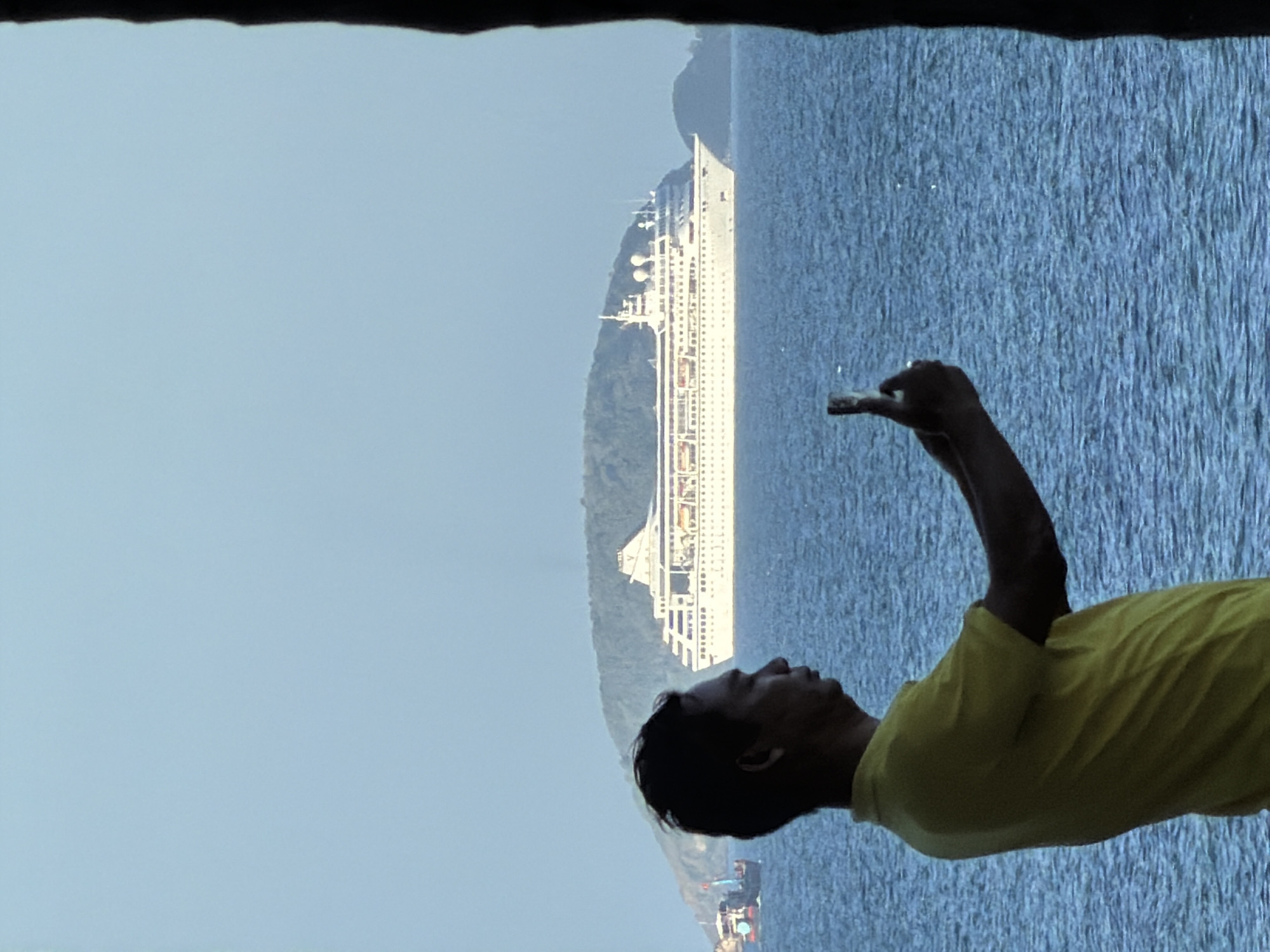 A man in a yellow shirt takes a photo with his phone of a large white cruise ship in the ocean, with a lush green island in the background. The sea is calm and blue, and the sky is a pale blue. A smaller boat is visible to the left of the cruise ship. The scene appears to be in Nha Trang, Vietnam, a coastal city known for its beaches and islands.