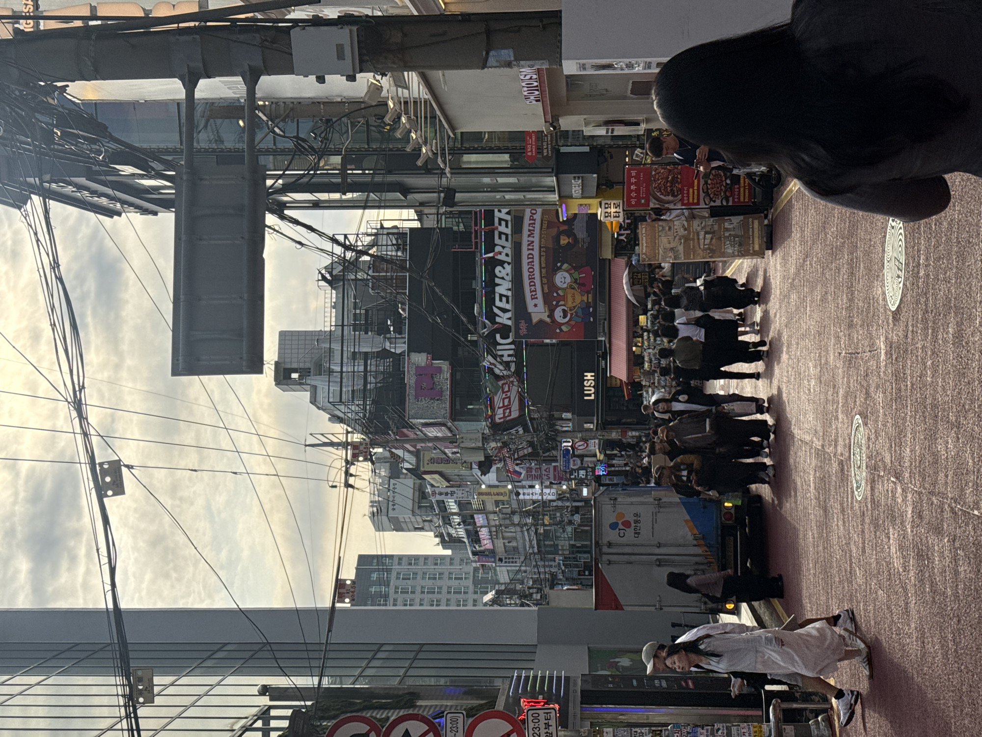 The image captures a vibrant street scene in Seoul, South Korea. The street is densely packed with signs and businesses. A group of people walks down the center of the street, moving away from the camera. A truck is partially visible near the group, and a couple is walking toward the camera on the right side of the image. Overhead, a dense network of wires stretches across the street. Signage indicates the location is likely "Redroad in Mapo".