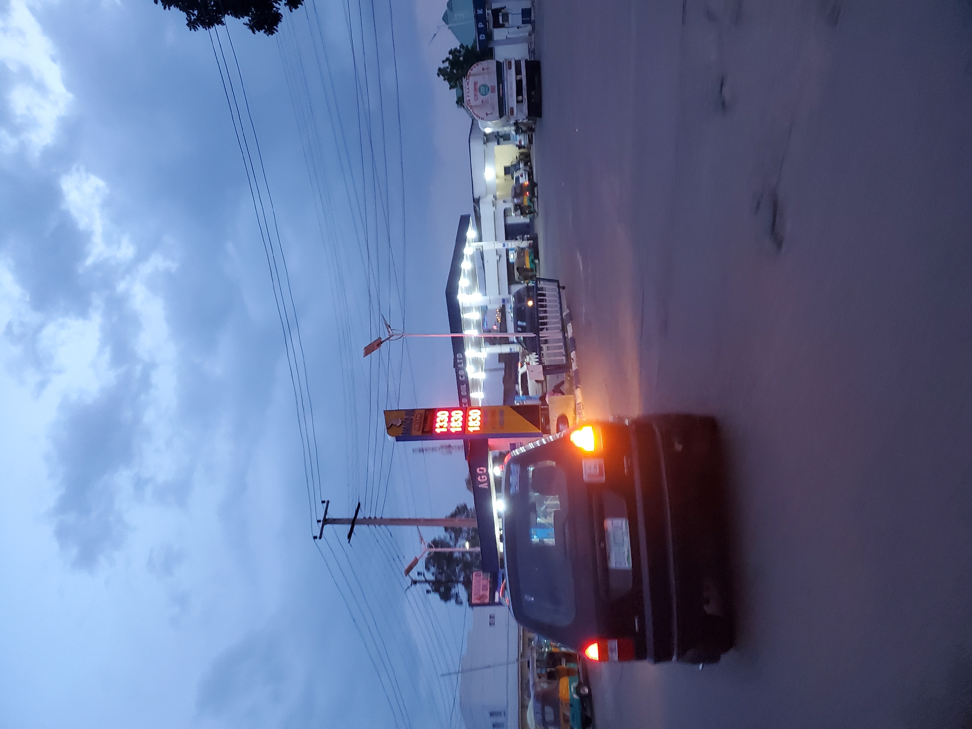 In Jos, Nigeria, at dusk, a dark-colored SUV with illuminated taillights is captured in motion on an asphalt road. The vehicle is in the foreground, with its rear facing the camera. The scene depicts a bustling gas station, identifiable by its illuminated pumps displaying prices like "1330," "1830," and "1830." A sign above the pumps reads "AGCO OIL CO. LTD." Several other vehicles, including a white tanker truck and what appear to be auto-rickshaws, are present at the gas station. The sky is overcast with a blend of blue and gray clouds, indicating the late hour. Overhead power lines stretch across the frame, supported by wooden poles. The overall environment suggests a typical urban setting in Jos, Nigeria, during the twilight hours.