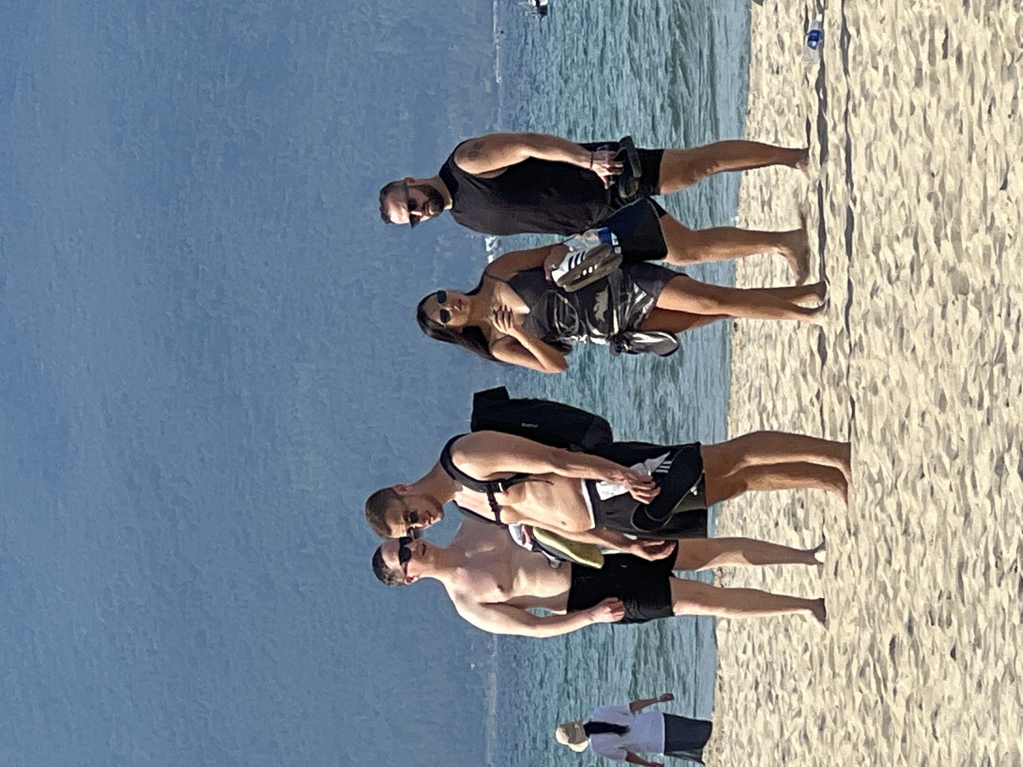 The image depicts a group of people on a sandy beach in Đà Nẵng, Vietnam. In the foreground, four individuals are visible. Two men, shirtless and wearing black swim trunks, stand close together, one with a black backpack. One man is holding a pair of flip-flops. Next to them, a woman in a black dress and a man in a black sleeveless shirt and black shorts are also present. The woman is holding a pair of flip-flops. In the background, a person in a white shirt and dark skirt is walking away from the camera. The ocean, with gentle waves and distant boats, stretches behind the beach, and a mountain range is visible on the horizon under a clear blue sky.