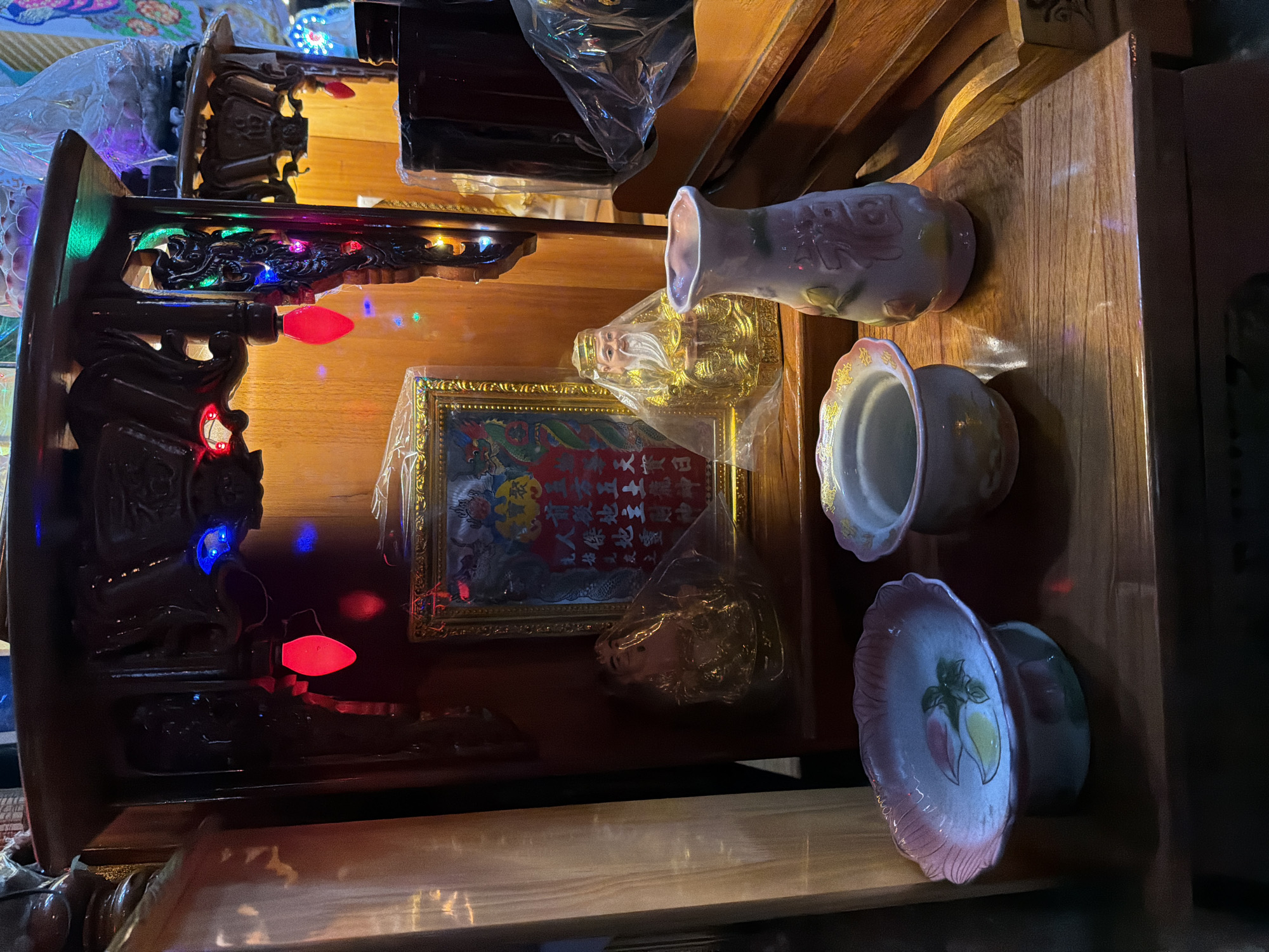 The image shows an indoor altar dedicated to deities, likely in a residential or commercial space in Nha Trang Ward, Vietnam. The altar is constructed of dark wood and is adorned with red, green, and blue fairy lights. Central to the altar is a framed painting depicting figures and Chinese characters, possibly a religious scripture or invocation. To the left of the painting is a small golden statue of a seated figure, draped in plastic. To the right of the painting is another golden statue, this one of an elderly man with a long white beard, also covered in plastic. In front of the altar, arranged on a wooden surface, are three ceramic vessels. The leftmost is a shallow, pink-rimmed dish decorated with fruit motifs. Next to it is a small, white, open-top pot with a decorative rim. To the right is a light pink vase with a floral design and Chinese characters. The overall impression is of a shrine or devotional area, possibly a place of worship or a household altar.