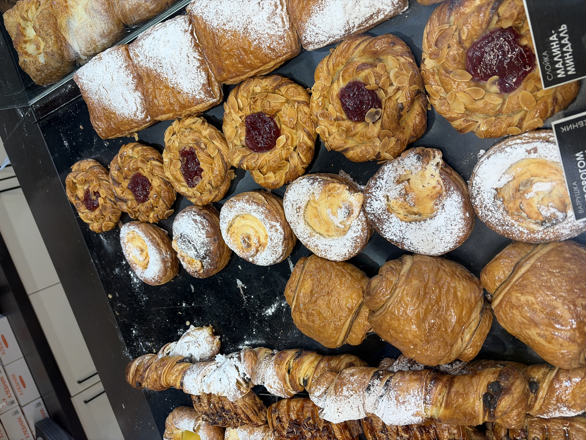 This high-angle, close-up shot captures a vibrant display of various baked goods, likely in a bakery or supermarket in Saint Petersburg, Russia. The scene showcases an abundant selection of pastries arranged on a dark, reflective counter, with some items appearing under a clear glass or plastic barrier.
The subjects are predominantly sweet pastries of different shapes and fillings. In the background, several rectangular, golden-brown pastries dusted with powdered sugar are visible. The mid-ground features round, spiraled pastries topped with flaked almonds and a generous dollop of red jam, possibly raspberry, also some partially covered in powdered sugar. There are also smaller, simpler round pastries, some plain, some dusted. In the foreground, larger, golden-brown, folded pastries, reminiscent of *pain au chocolat* or similar laminated dough items, are present. Below these, twisted pastries with visible dark fillings, possibly chocolate or poppy seeds, are arranged, some dusted with powdered sugar.
The activity depicted is the presentation of freshly baked goods for sale. The arrangement suggests a well-stocked and inviting bakery display.
Notable details include the texture and shine of the pastries, indicating freshness. Several items are artfully decorated with powdered sugar, flaked almonds, and fruit jam.
Visible text on black labels provides identification: one label reads "СЛОЙКА МАЛИНА-МИНДАЛЬ" (Sloyka Malina-Mindal), meaning "Puff pastry raspberry-almond." Another partially visible label mentions "ВАТРУШКА... ТВОРОГОМ" (Vatrushka... Tvorogom), indicating a sweet bun with cottage cheese. On the far left, multiple white boxes are visible with the Russian word "ХЛЕБНИК" (Khlebnik), which translates to "baker" or "bread maker." The lighting is bright and even, typical of an indoor commercial setting.