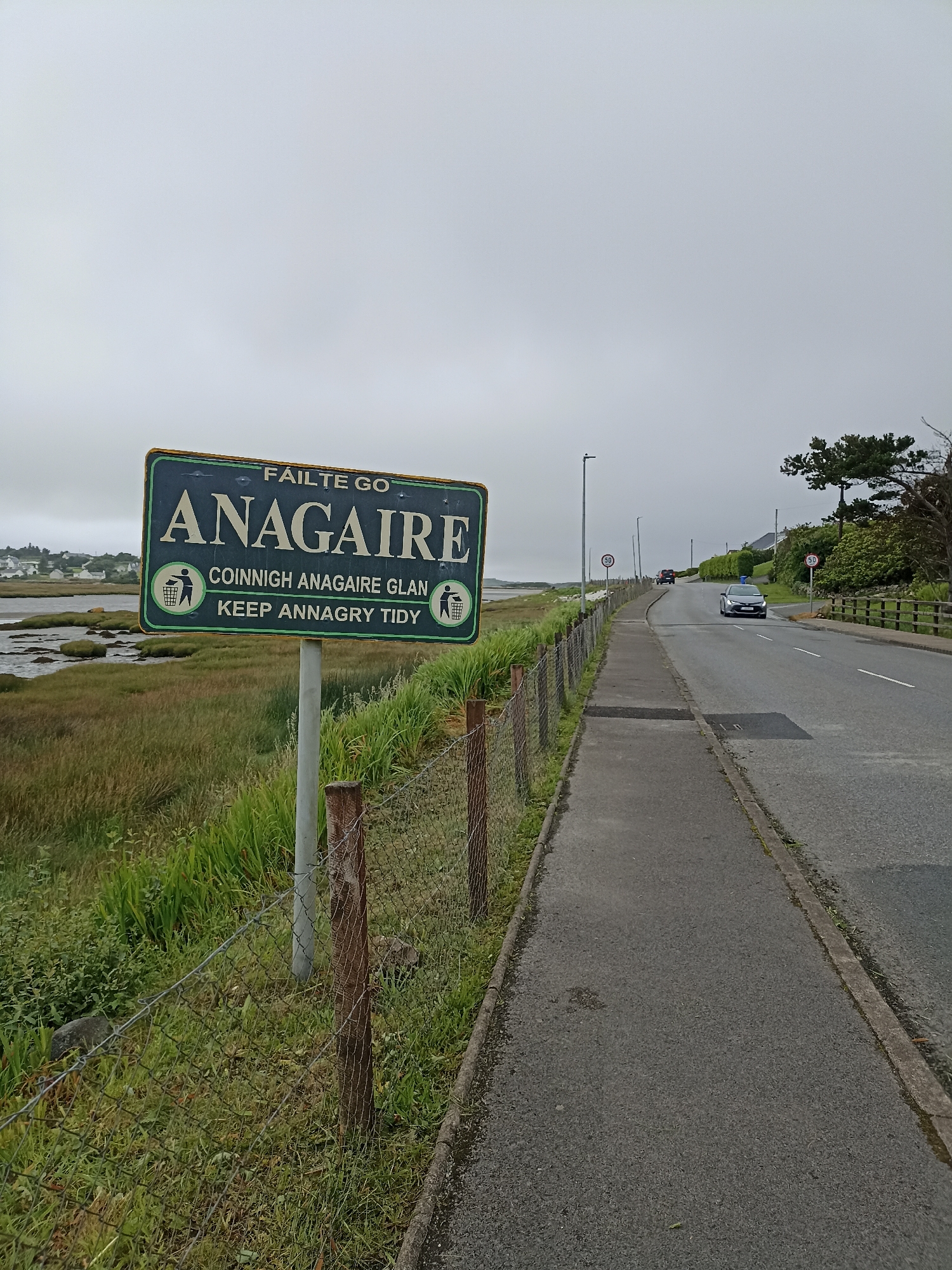 The image depicts a scene in Annagry, County Donegal, Ireland, on an overcast day. A green sign with gold lettering is prominently displayed in the foreground, reading "FÁILTE GO ANAGAIRE" and below that, "COINNIGH ANAGAIRE GLAN KEEP ANNAGRY TIDY," accompanied by two icons of a person discarding litter into a bin. The sign is mounted on a metal pole and situated beside a chain-link fence that runs along a grassy verge. Beyond the fence, a body of water with marshy vegetation is visible, with a small village or town in the distance. To the right of the sign, a paved road with a sidewalk runs alongside the fence. Several cars are visible on the road, including one in the middle ground approaching the viewer and another in the distance. Speed limit signs indicating "50" are present on poles along the road. Trees and shrubbery line the far side of the road, with a wooden fence running parallel to it. The overall environment suggests a coastal area with some residential development.