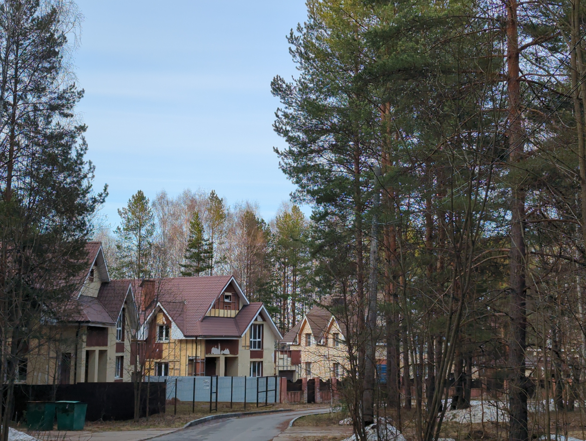 The image depicts a residential area in what appears to be a suburb of Chaikovsky, Russia. Several houses are visible, some under construction or renovation, with exposed brickwork and incomplete facades. A gray metal fence partially encloses at least one of the properties. The road in front of the houses is paved and slightly winding. Trees, predominantly pines and deciduous varieties without leaves, dominate the landscape, bordering the street and houses. Patches of snow are visible on the ground, suggesting a late winter or early spring setting. Two green dumpsters are near the beginning of the road. The sky is pale blue with thin clouds.