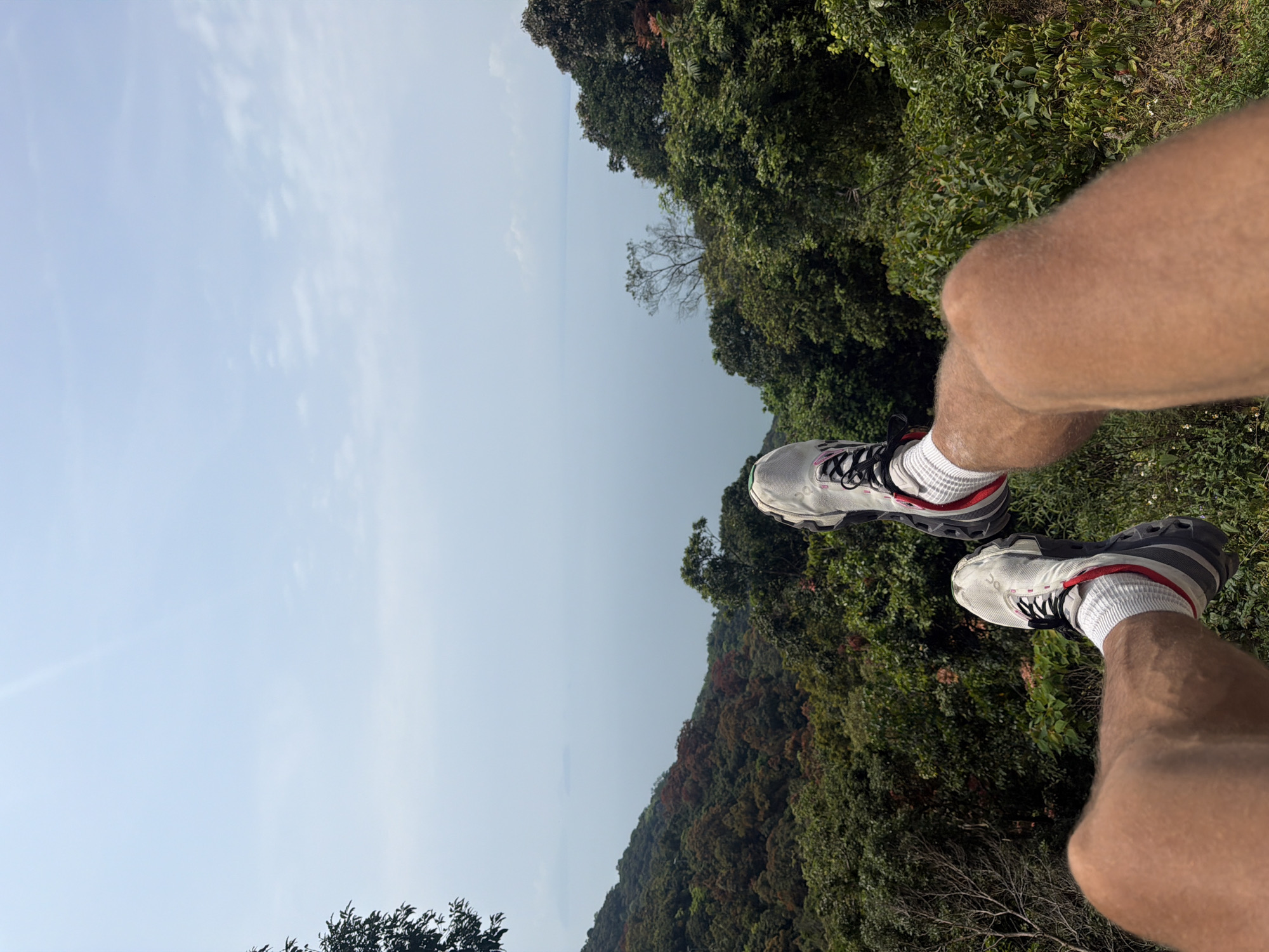 The image shows a first-person view of a person sitting with their legs dangling over a forested hillside. The sky is visible in the background. The person is wearing white and gray running shoes with red accents and white socks. The hillside is covered in dense green vegetation, possibly a tropical forest. The location is possibly somewhere in the hilly region of Đà Nẵng, Vietnam.