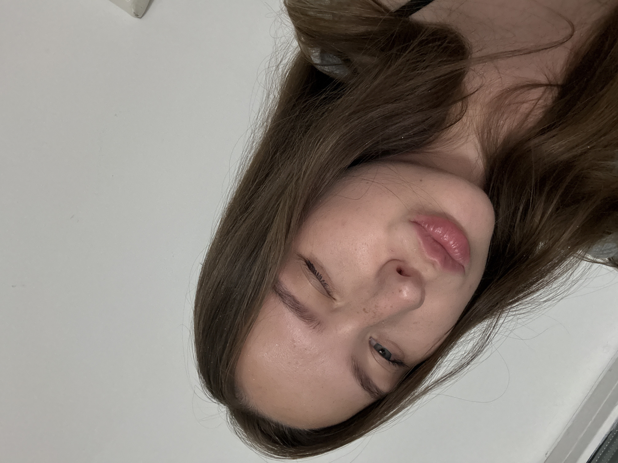 A young woman with long, brown hair is shown in a close-up, slightly angled selfie. She is winking with her right eye and puckering her lips. Her skin has a light dusting of freckles. The background is a plain, light-colored wall. There is no indication of the city of Chaikovsky, Russia, in the image.