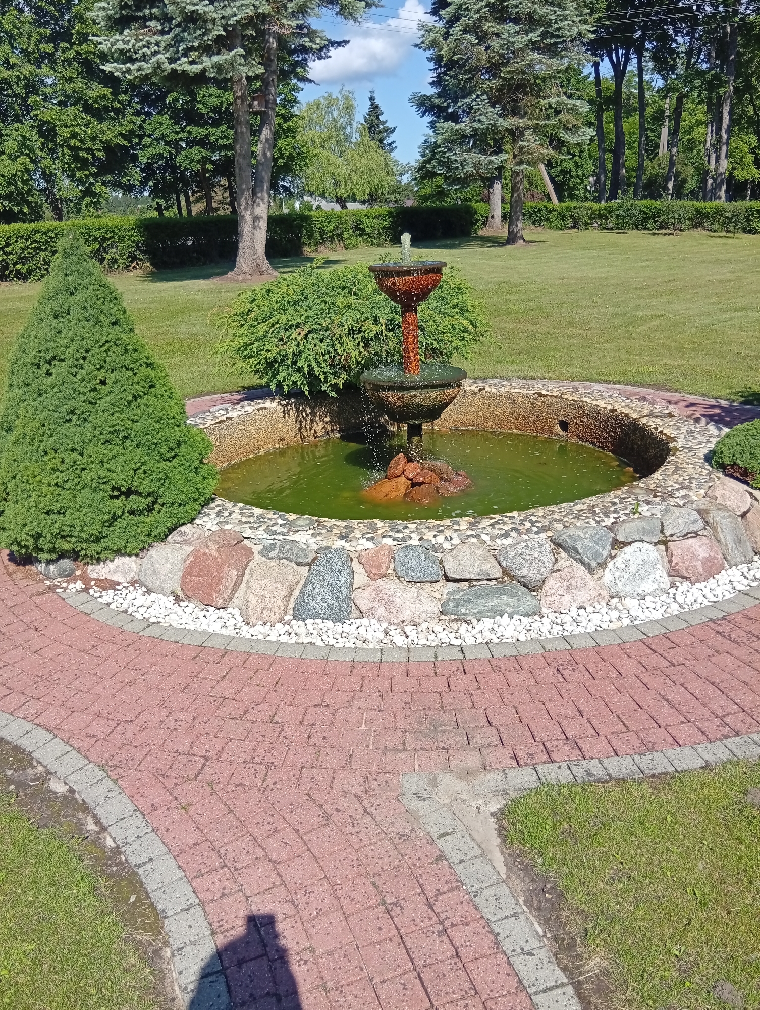 The image shows a garden area with a fountain as the central feature. The fountain is made of a reddish-brown material and has water flowing from its top tier. The fountain is situated in a circular pond with a stone border. Surrounding the pond is a pathway made of red brick pavers, bordered by smaller grey pavers and white gravel. A green, cone-shaped shrub is planted to the left of the fountain, and a larger, bushy green shrub is behind it. The background consists of a neatly manicured green lawn, several mature trees, and a line of trimmed hedges. The sky is visible above the trees, with some white clouds against a blue background. The scene appears to be an outdoor garden in a well-maintained residential or public space. The image is taken from a slightly elevated angle, looking down towards the fountain and pathway. The time of day appears to be daytime, with clear sunlight casting shadows, including a distinct shadow of a person in the foreground on the pathway. The location is stated to be Dirvonėnai, Lithuania.