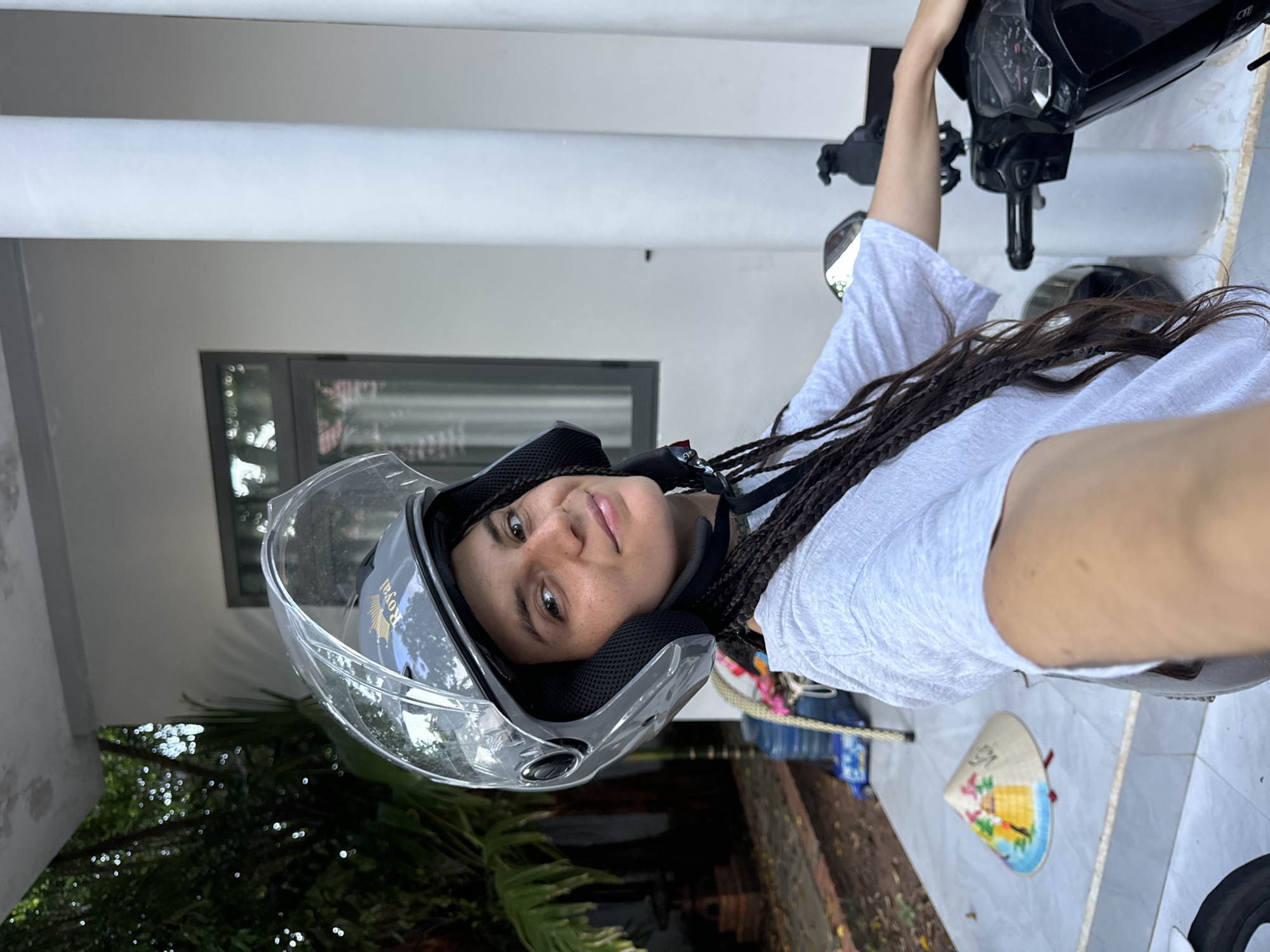A young woman with long, dark braided hair, wearing a grey t-shirt and a grey helmet with a clear visor, is seated on a motorcycle. She is looking towards the camera with a slight smile. The background shows a white building with large white pillars and a grey-framed window. To the left, there is lush greenery and trees. On the ground in front of the motorcycle, a conical Asian hat with colorful decorations is visible. The scene appears to be outdoors, possibly in a residential or roadside area in Mũi Né, Vietnam, given the presence of the conical hat.