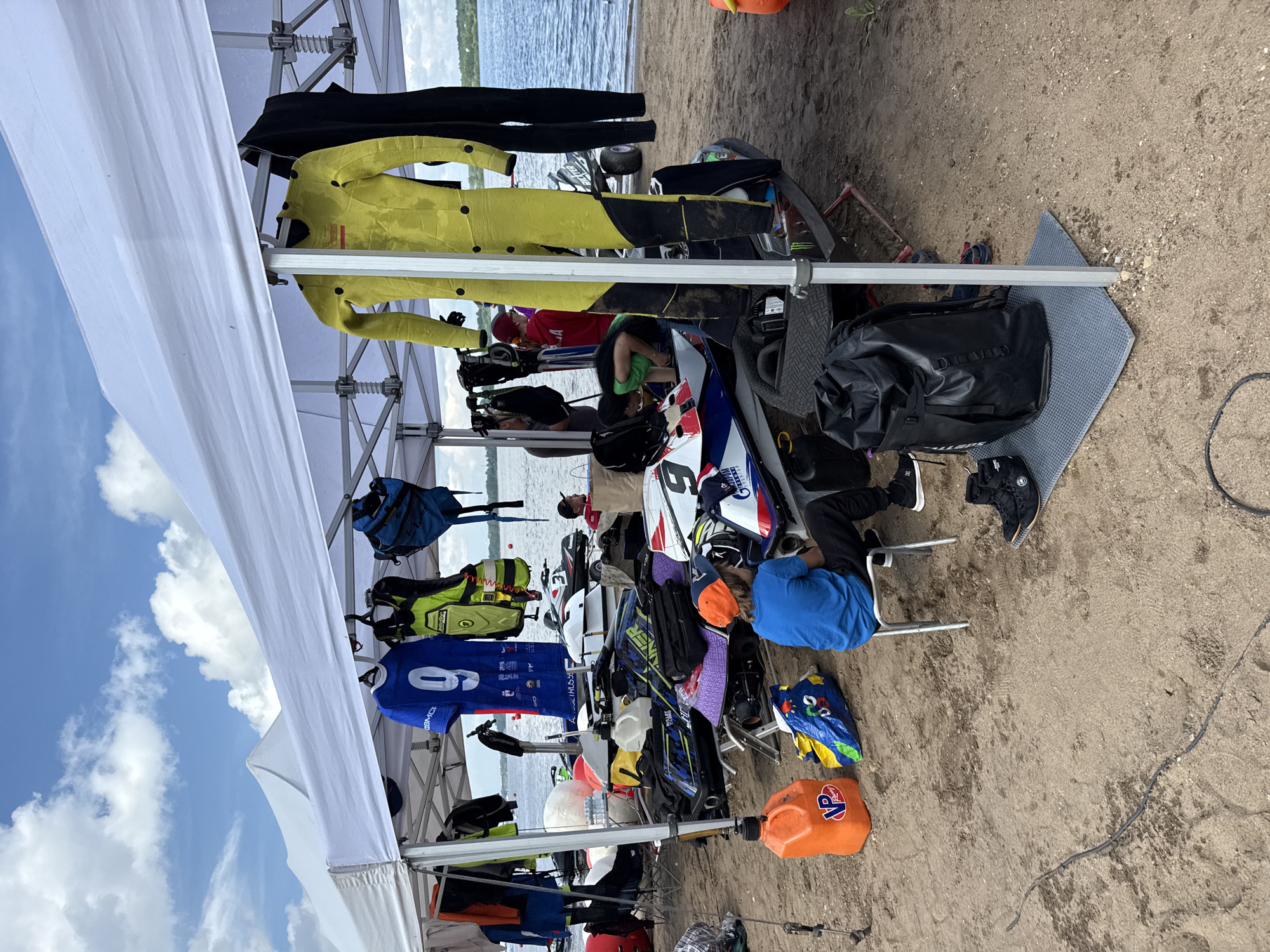A group of people are gathered on a sandy beach, likely for a watercraft event, under a white tent. Several jet skis are parked behind the tent. On the left side of the tent, an orange fuel can and a colorful bag are visible. A person in a blue shirt and orange cap is sitting on a small chair. A large black waterproof backpack rests on a mat in front of the tent. In the background, the blue water of a lake or river stretches out under a partly cloudy sky. The overall scene suggests a preparation or staging area for a recreational activity. The location is stated to be the city of Perm, Russia.