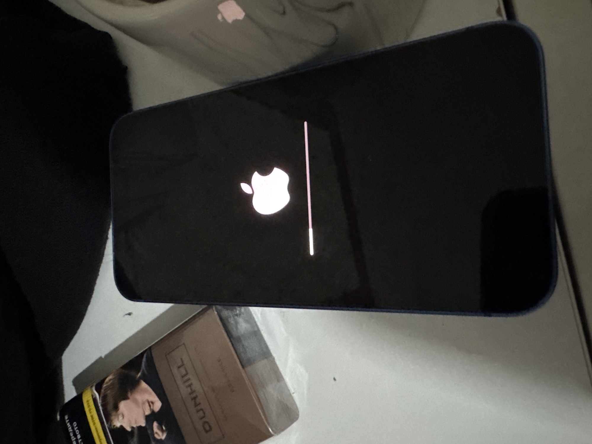 The image shows a black iPhone displaying the Apple logo and a loading bar on its screen. The phone is placed on a white surface, next to a pack of Dunhill cigarettes. The location cannot be determined from the image, so it cannot be stated to be Sofia, Bulgaria.