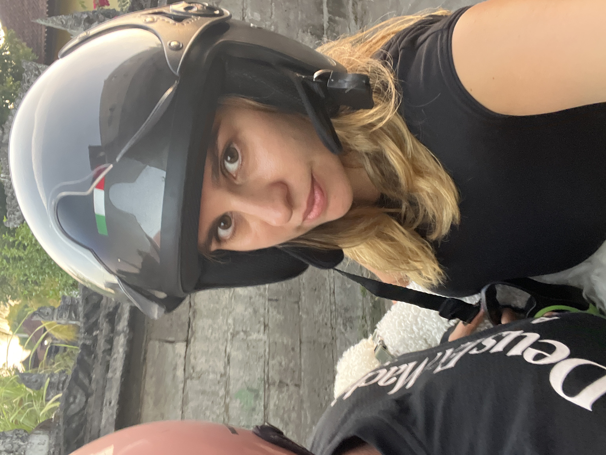 This is a selfie taken from a motorcycle in the city of Kediri, Indonesia. A woman with blonde hair and brown eyes is wearing a dark grey helmet with an Italian flag sticker on the front. She is looking at the camera with a slight smile. Beside her, on the left, is another person wearing a pink helmet and a black t-shirt with white lettering that appears to say "Deus Ex Machina." A white fluffy object, possibly a small dog, is visible between the two people. The background consists of a stone wall with some greenery and sunlight filtering through.