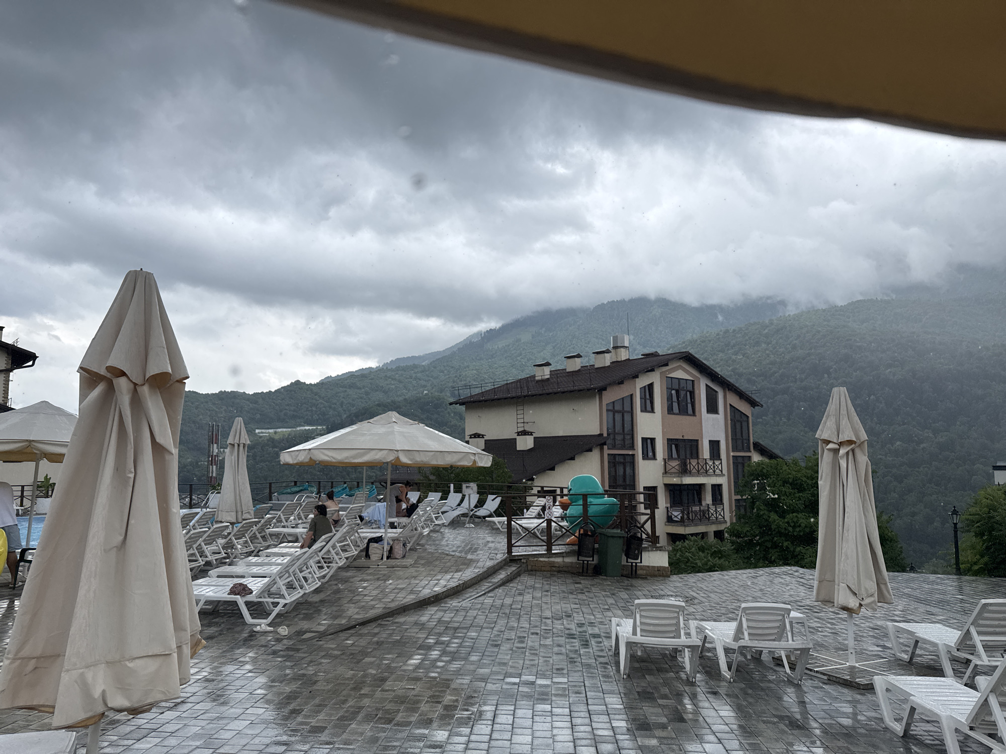 This image depicts a resort area in Krasnaya Polyana, Russia, under overcast and rainy conditions. The foreground is dominated by a wet, tiled patio with several empty white lounge chairs. A large, closed cream-colored umbrella stands prominently on the left. In the middle ground, more lounge chairs are arranged, some occupied by a few individuals. A beige umbrella is open over a section of these chairs. Behind the lounge chairs, a multi-story building with balconies is visible, situated against a backdrop of lush green, forested mountains shrouded in mist. A large, teal-colored inflatable duck is positioned on a deck area. The sky is heavily clouded, suggesting continued inclement weather.