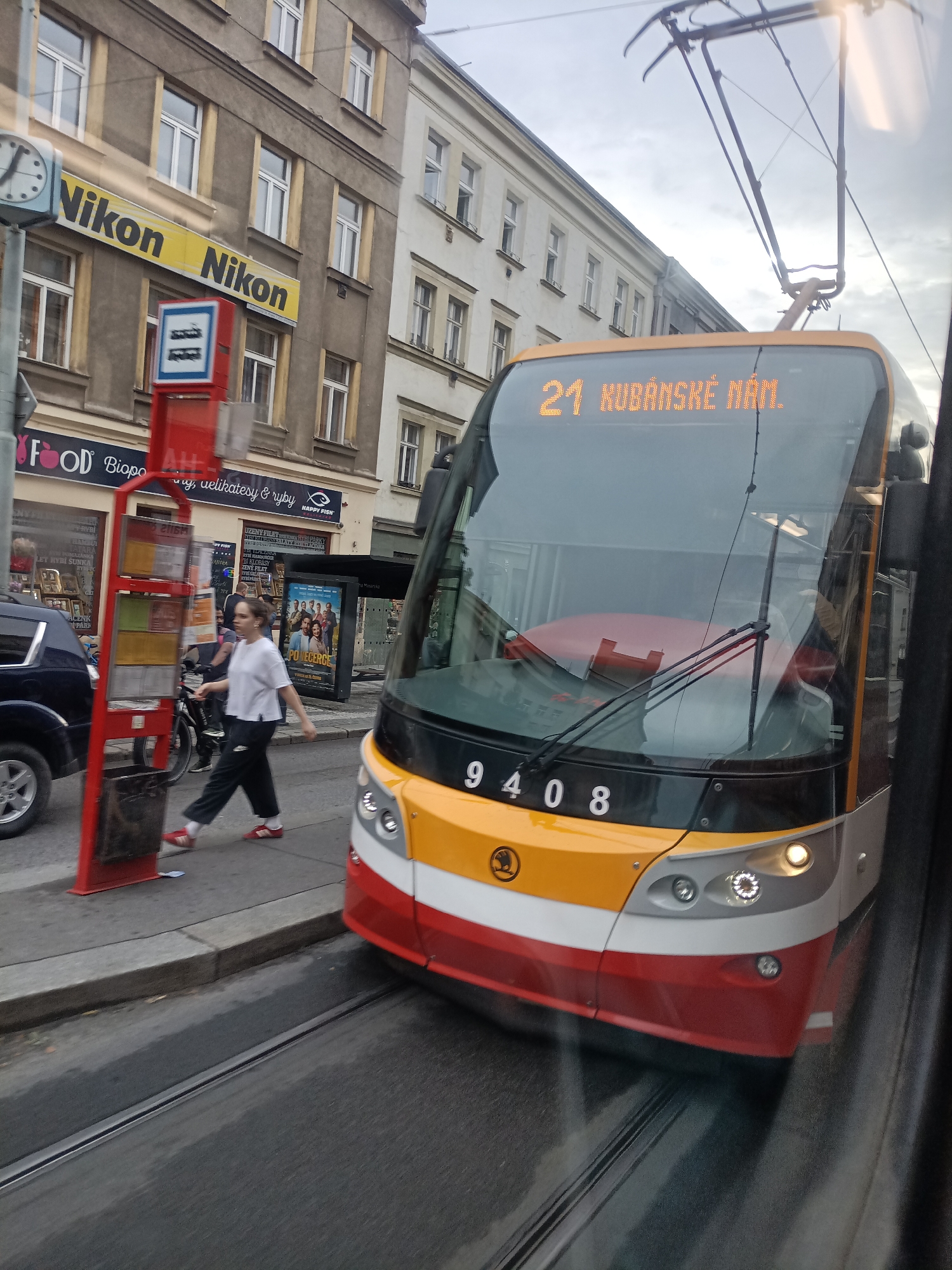 This media file captures a street scene in Prague, Czechia. The primary subject is a yellow, white, and red tram, numbered 9408, moving forward and slightly to the right. The tram displays "21 KUBÁNSKÉ NÁM." on its front, indicating its route number and destination.
In the foreground, a young woman with dark hair, wearing a white t-shirt and black athletic pants with red sneakers, is walking across a street, passing by a red bus stop shelter. She appears to be focused on her movement. Behind her, a bicycle is visible. To the left of the woman, a dark blue car is parked at the curb.
The background features buildings with multiple windows, typical of urban architecture. A large yellow sign with black lettering reading "Nikon Nikon" is prominently displayed on one of the buildings, alongside a clock face. Below the Nikon signs, shop fronts are visible, one displaying "FOOD Biopo" and another with "HAPPY FISH" and text in Czech, including "UZENY FILETRYBLPARKY". A movie poster, with the title "POVECERCE", is also visible.
The scene appears to be set during the daytime, possibly late afternoon, given the lighting and the overcast sky. The overall atmosphere is that of a bustling city street. The tram's active presence suggests it is in motion, carrying passengers. The focus on the tram and the street activity creates a sense of urban movement and daily life.