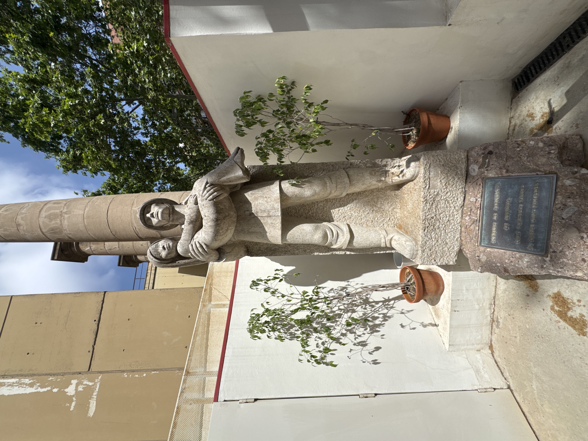 The image features a stone statue depicting two figures, an adult and a smaller person, set within an urban environment. The larger figure, standing upright, is shown cradling or carrying the smaller, prostrate figure in their arms. Both are carved in a robust, somewhat blocky style from light-colored stone, emphasizing strength and endurance. The statue is positioned on a textured stone base, which forms part of a larger architectural structure.
On the right side of the statue's base, a rectangular bronze plaque is affixed. The inscription, in Catalan, reads:
"BOMBERS DE BARCELONA" (Firefighters of Barcelona)
"EN MEMÒRIA ALS QUE DONAREN" (In memory of those who gave)
"LA SEVA VIDA EN COMPLIMENT" (Their life in fulfillment)
"DEL DEURE" (Of duty)
"ALS SALVADORS DEL POBLE" (To the Saviors of the People)
This text identifies the statue as a memorial dedicated to the firefighters of Barcelona who died in the line of duty.
The scene is brightly lit by natural daylight, evident from the strong shadows cast on the white wall behind the statue and the ground. Two small potted plants with green leaves are placed near the statue, one on either side. In the background, a large tree with abundant green foliage is visible against a clear blue sky dotted with white clouds. A substantial fluted column stands behind the statue to the left. Portions of light-colored building walls, some showing signs of age, frame the setting. The overall impression is a solemn tribute in a public, sunlit area of Barcelona, Spain.