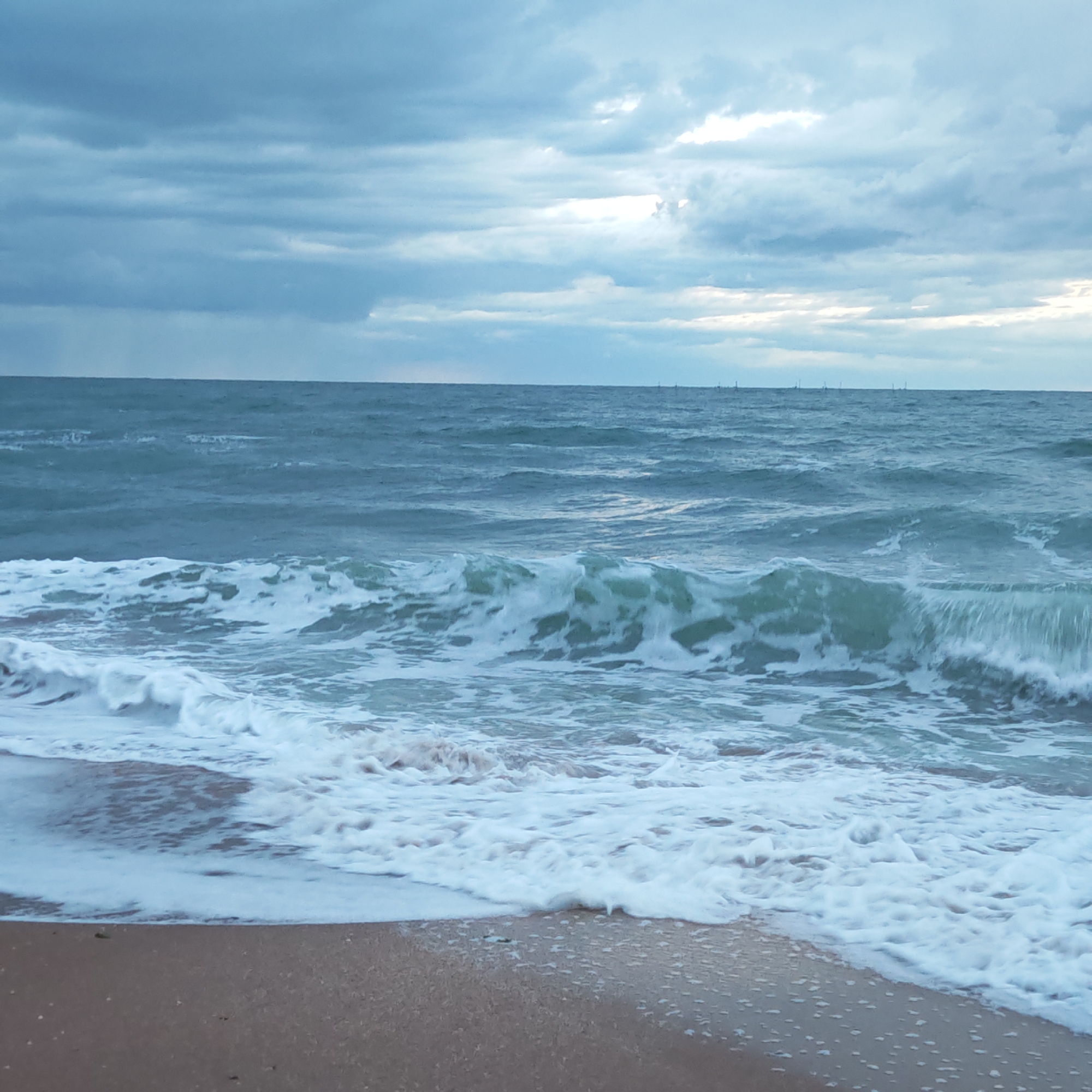 The image depicts a beach scene in городское поселение Щелкино, Ukraine. The foreground shows a sandy beach with wave foam reaching the shore. The sea stretches to the horizon under a cloudy sky. Distant, faint vertical structures are visible on the horizon line, possibly sailboats or distant coastal structures. A wave is breaking in the middle ground with sea foam.