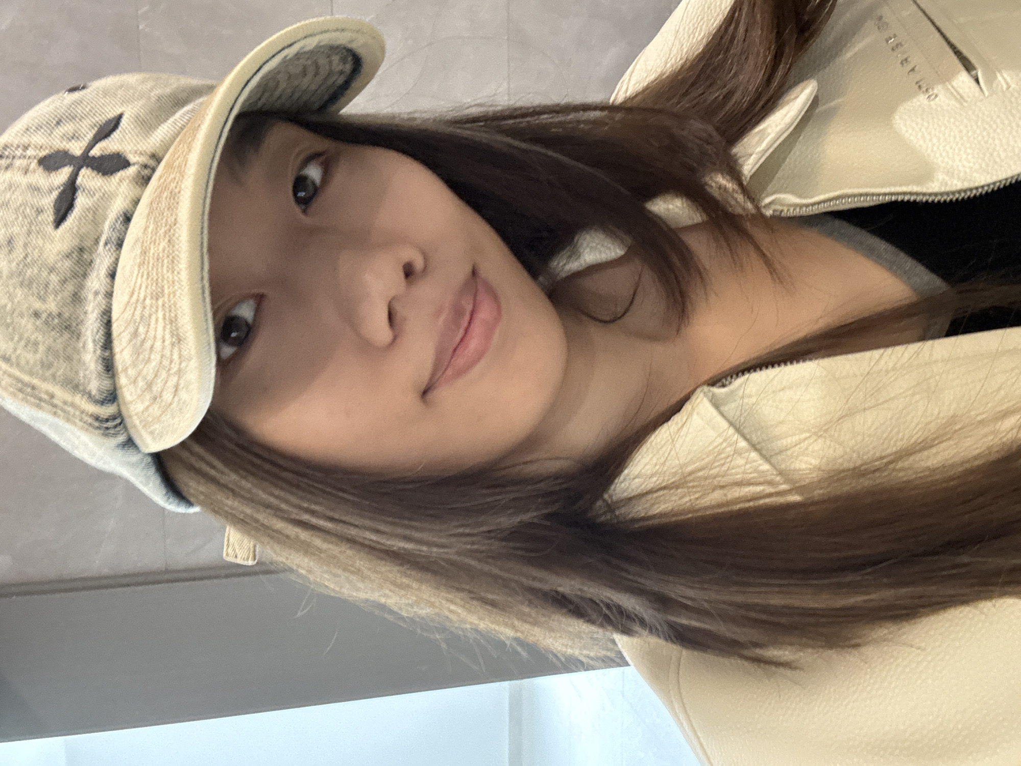 A young woman is posing for a photo. She is wearing a light-colored denim hat with a dark "X" on the front. She has long, dark brown hair and is wearing a light-colored leather jacket. The background appears to be a wall with a grey and white tile pattern. There is no discernible indication of the location being in Nan'an District, China.