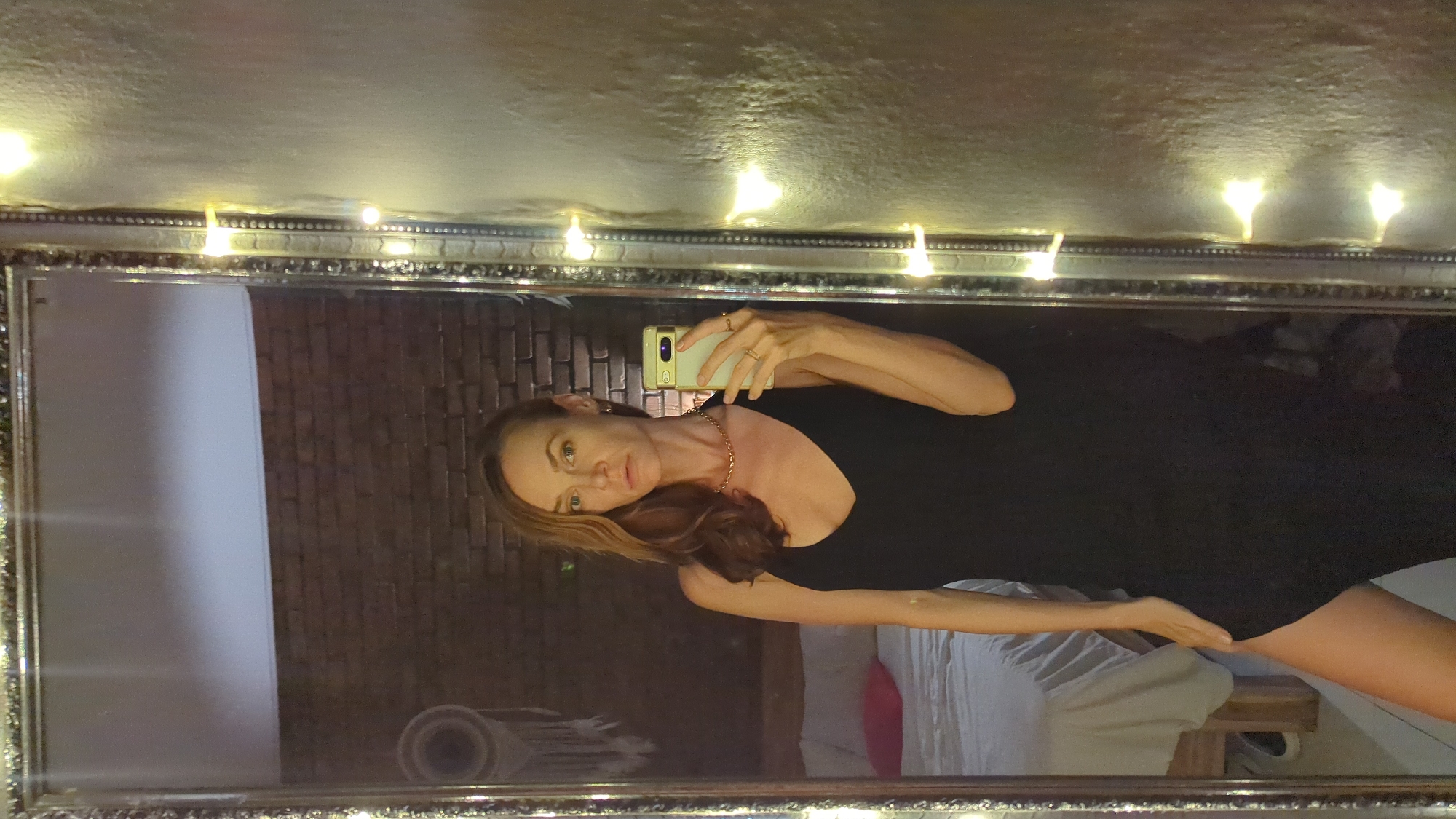 A mirror selfie captures a woman standing in a room, likely a bedroom, in North Kuta, Indonesia. She is the central subject, looking directly into the camera of a light-colored smartphone (possibly a Google Pixel) that she holds up to take the photo. She wears a simple black top or dress, a gold chain necklace, and rings on her fingers. Her long, light brown hair is styled down, partially covering her shoulder.
The large mirror, framed with ornate silver-colored detailing, reflects the room. Along the top edge of the mirror's frame and on the wall above it, several glowing string lights (fairy lights) are draped, providing warm illumination and creating a cozy, evening ambiance. The wall where the mirror hangs appears to be textured and light-colored.
In the reflection behind the woman, a rustic exposed brick wall is visible, adding an industrial or earthy element to the decor. To the right of the reflection, part of a bed with white bedding and a glimpse of a pink pillow can be seen. On the floor, near the bottom left of the reflection, a rolled-up mat, possibly a yoga mat, is visible. The overall scene suggests an indoor setting during the evening or night, relying on artificial light sources. No discernible text is visible in the image.