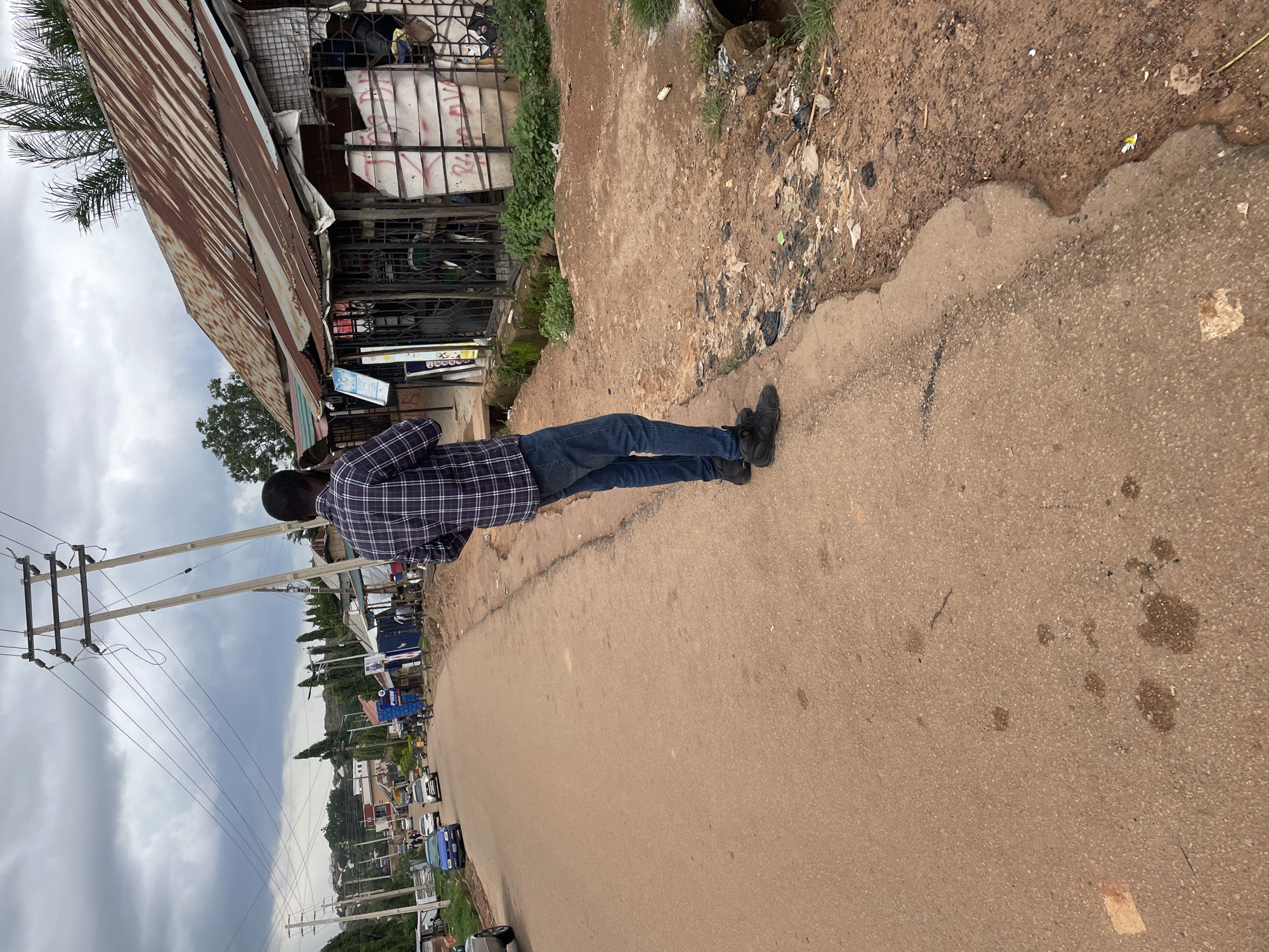 An over-the-shoulder, medium shot captures a person standing on the side of a dirt road, facing away from the camera. The person is wearing a plaid shirt and blue jeans and is holding a cell phone to their ear, appearing to be in a conversation. The environment is an urban setting, possibly in Jos, Nigeria, with a mix of dilapidated buildings and a busy street. A corrugated iron roof structure is visible to the right, with metal bars and graffiti on its facade. A narrow gutter runs along the side of the road, filled with debris and stagnant water. Utility poles with electrical wires stretch across the cloudy sky in the background. Cars and other vehicles are seen driving down the road, suggesting an active street scene. The ground is a mixture of red soil and asphalt, with scattered litter indicating a lack of proper waste management.