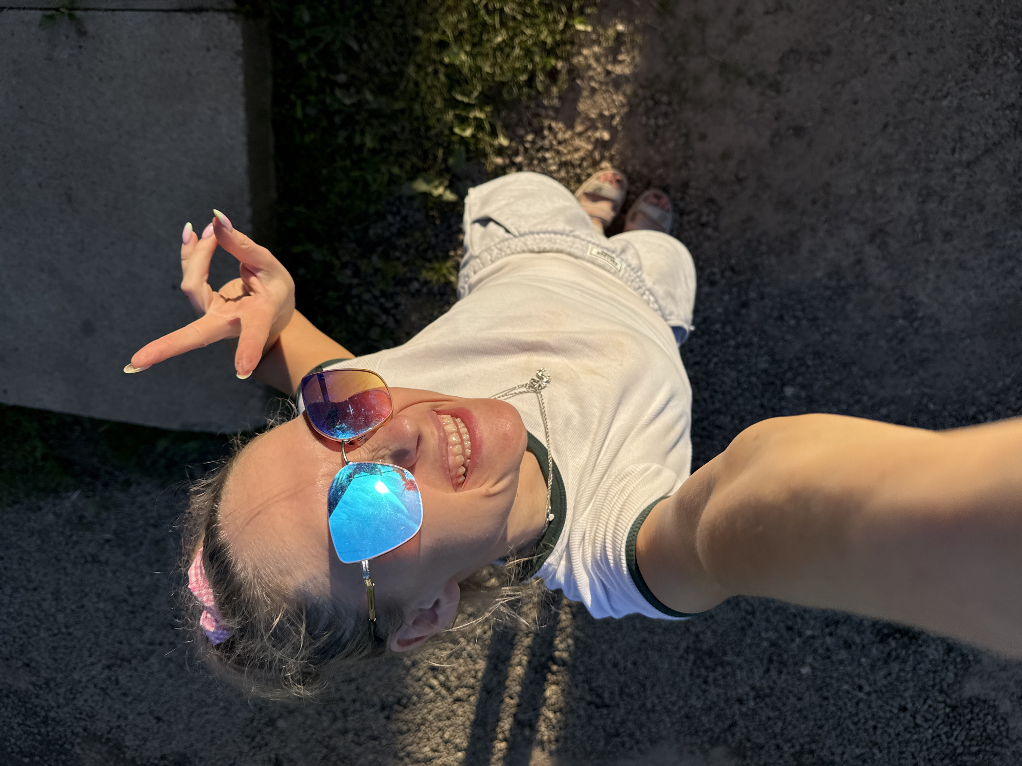A woman is taking a selfie outdoors in a rural or suburban area, possibly in the city of Fokino, Russia, given the context. She is wearing sunglasses with blue and rainbow lenses, a white t-shirt with a green trim around the neck, and light-colored shorts. Her hair is pulled back with a pink scrunchie, and she is smiling, showing her teeth. Her right hand is raised, making a peace sign. She is wearing sandals and standing on a gravelly surface. The lighting suggests it is either early morning or late afternoon, with sunlight casting shadows. The background consists of more gravel and some greenery, with a dark vertical structure to the right.