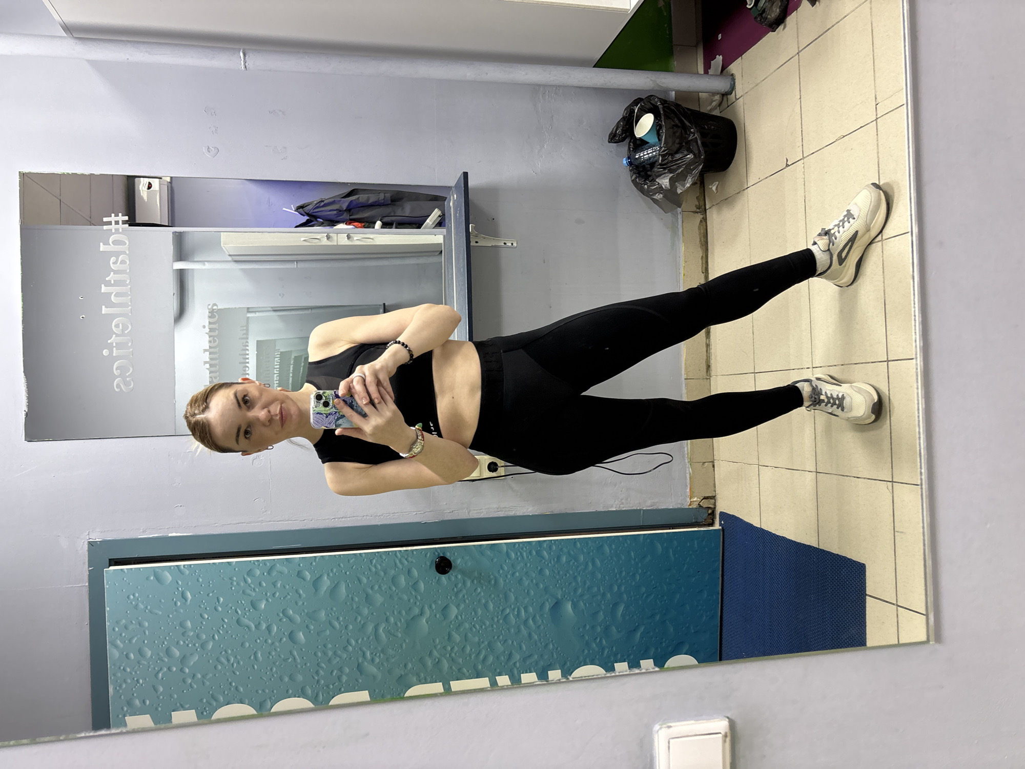 A young woman poses in front of a mirror, capturing a full-body selfie with her mobile phone. She is wearing black athletic attire: a sports bra and leggings. Her hair is pulled back. The reflection reveals a locker room environment, characterized by tiled walls and floor, with a trash bag hanging near the upper right corner. A blue panel with graphic water droplets is attached to the wall near the bottom of the frame. The reflected signage on the mirror reads "athletics #". There is no indication of a location in Saint Petersburg, Russia.