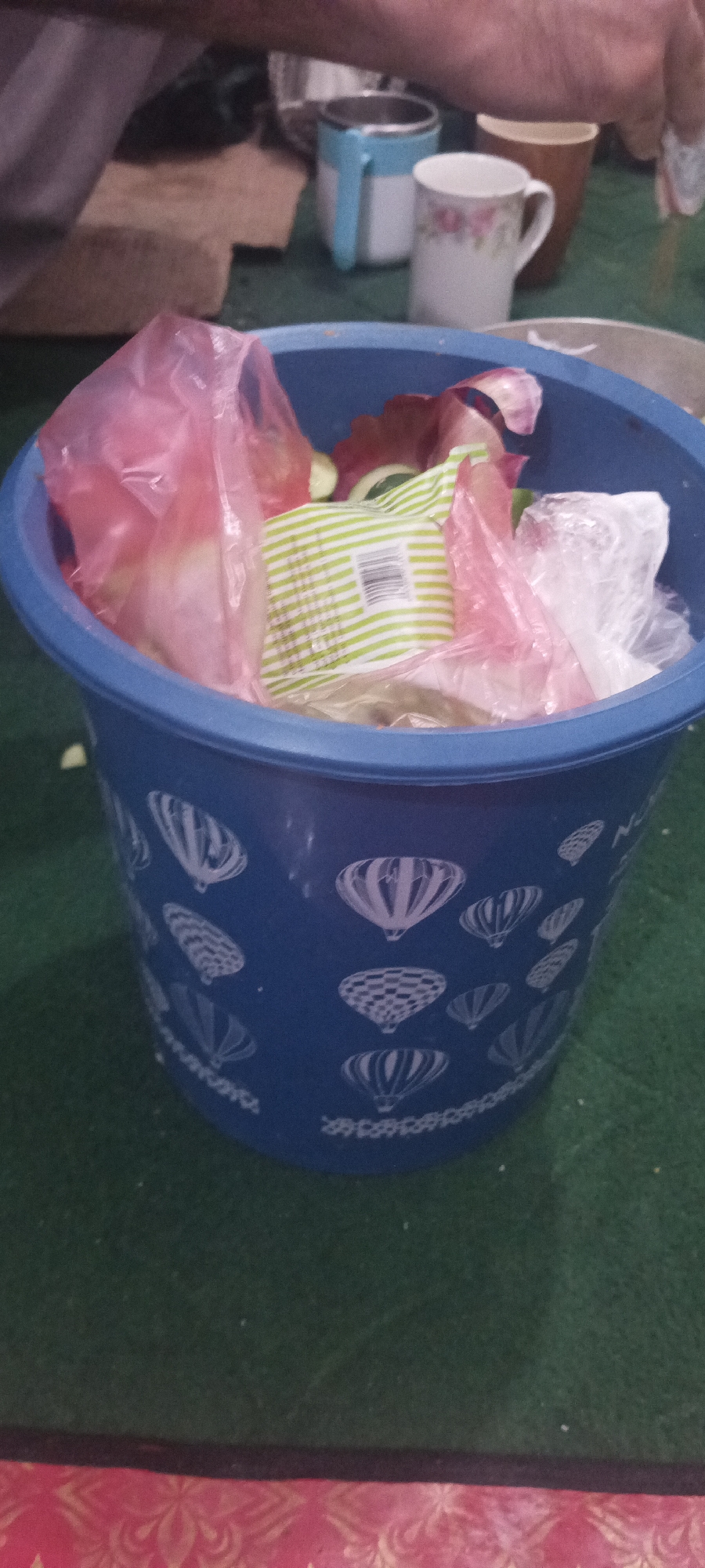 A blue plastic trash can, decorated with white images of hot air balloons, is overflowing with trash. The trash includes pink plastic bags, a green and white striped package, and what appears to be food scraps. The trash can is situated on a dark green surface, possibly carpet or a rug, with a pink and gold patterned fabric visible at the bottom of the frame. In the background, partially visible, are a person's arm and hand, a metal and blue mug, a white floral patterned mug, and a brown mug. The scene suggests a domestic or public setting where waste is being discarded.