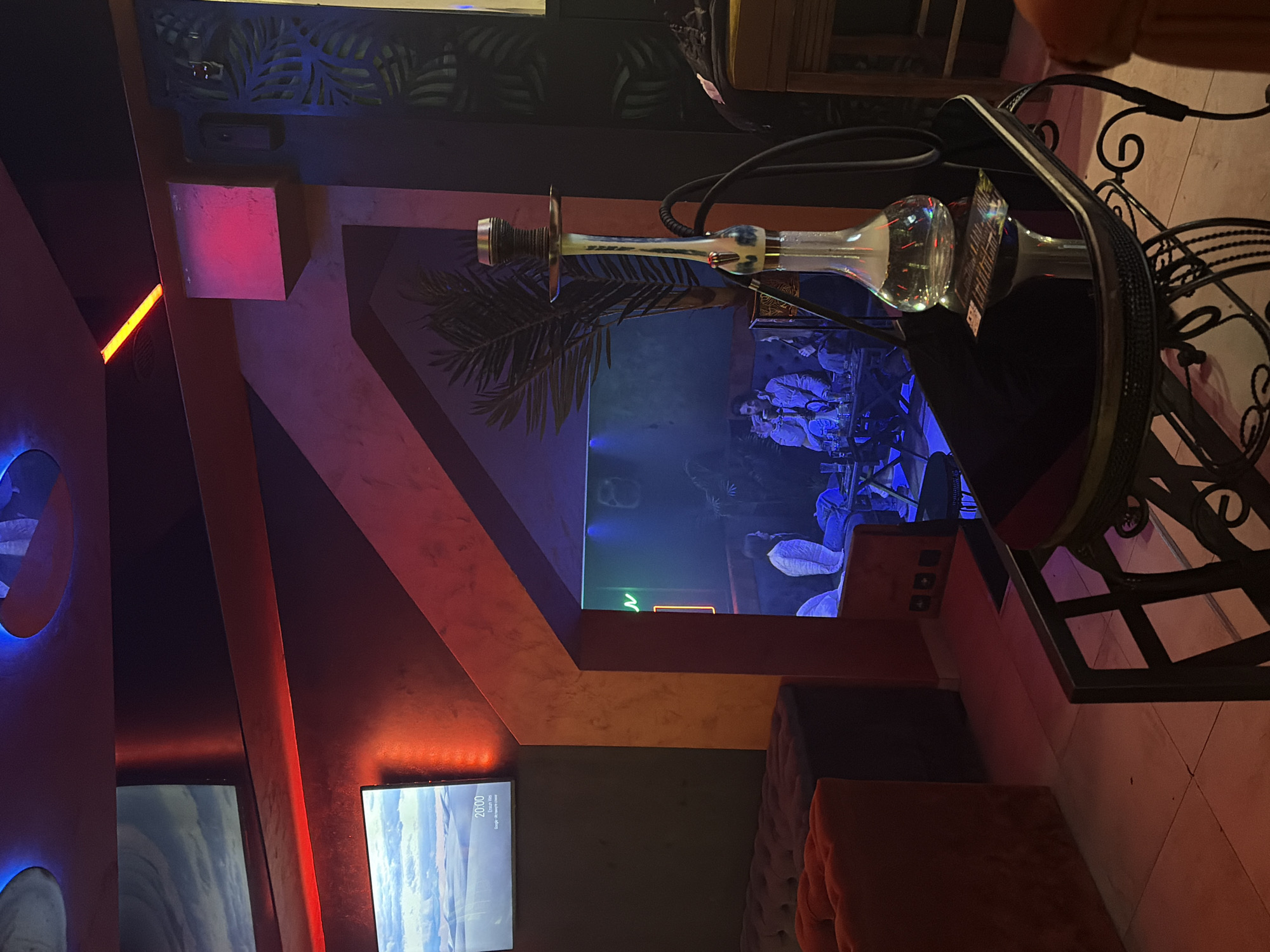 The image captures an indoor scene in Budva, Montenegro. A hookah sits on a black table with ornate metal legs and frame. The table also has a black case on top. In the background, a palm tree design is painted on the wall, and a video screen displays a scene with several people in blue lighting. Another TV screen is visible in the lower-left corner. There are geometric wall decorations with red, pink, and blue lighting, and cushioned furniture, partly visible in the foreground.