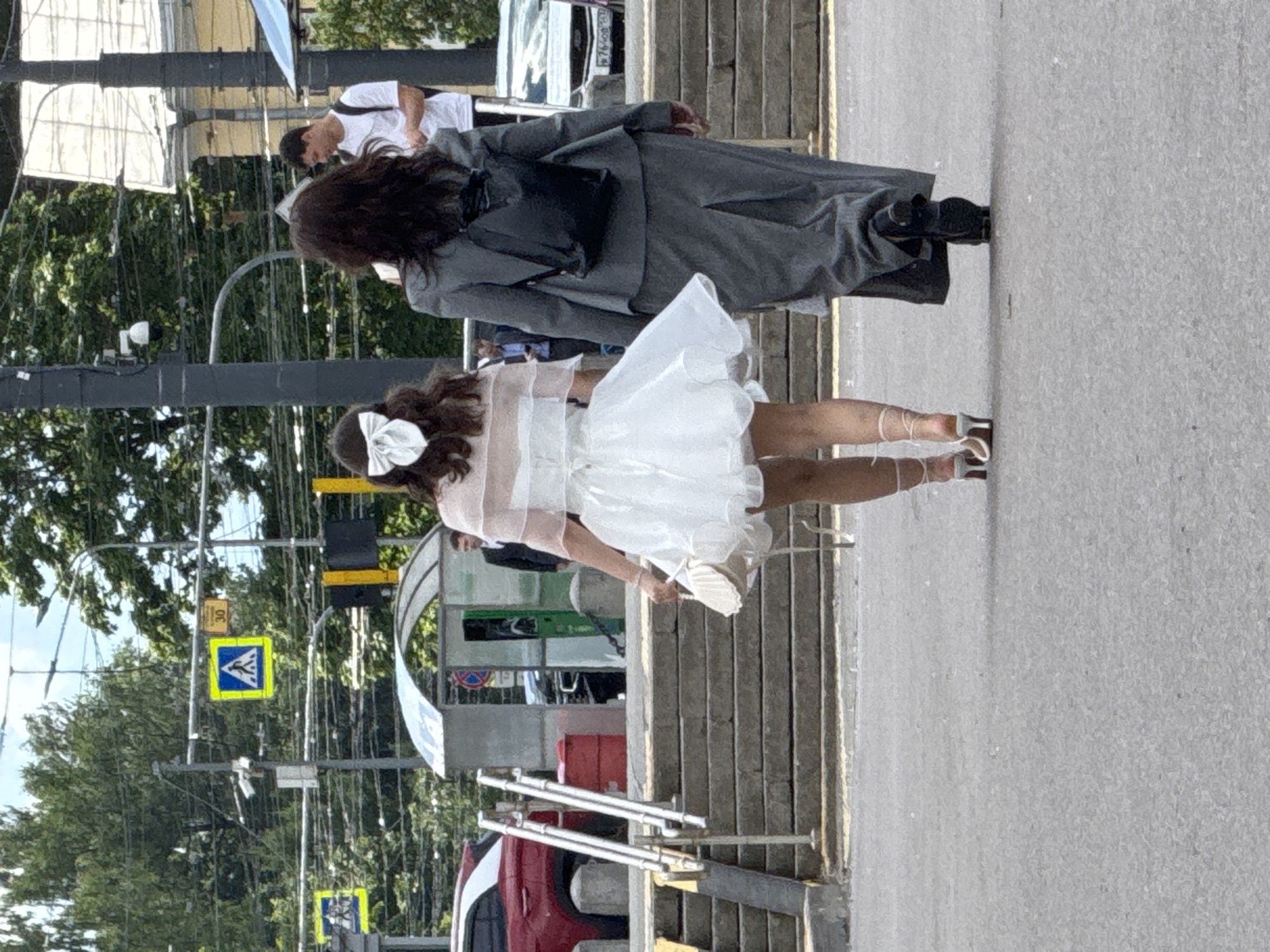 The image depicts two individuals outdoors in what appears to be a city setting. The primary subject in the foreground is a person wearing a short, white, layered dress with a large white bow in their hair. They are also wearing white heeled sandals with straps that tie around their ankles. This person is holding a pair of white shoes in their right hand. Behind them, another individual is visible from the waist up, wearing a long, dark grey coat over a dark garment. They have long, dark, wavy hair.
The background suggests an urban environment. There are elements of street infrastructure, including traffic lights, street signs, and what looks like a pedestrian crossing. Trees with green foliage are visible, indicating it is likely daytime during a warmer season. The ground surface appears to be a paved sidewalk or street, with wooden steps or a ramp also present. In the distance, parts of buildings and vehicles are partially visible.
The orientation of the image is rotated, making it appear sideways. The lighting suggests it is daytime, possibly sunny, given the shadows. The overall mood is ambiguous, as the individuals' expressions are not clearly visible due to their orientation and the image resolution.
One visible sign is a yellow square sign with a blue pedestrian symbol. Another yellow sign with the number "30" is also present. The location is identified as Saint Petersburg, Russia. The activity appears to be casual, with the individuals walking or standing on a street.